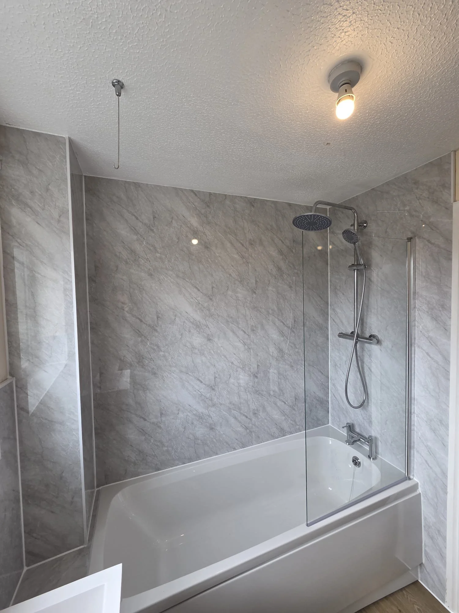 A modern bathroom with a bathtub, a glass shower wall, a rainfall showerhead, a hand-held showerhead, a textured ceiling with a light bulb, and gray marble walls.