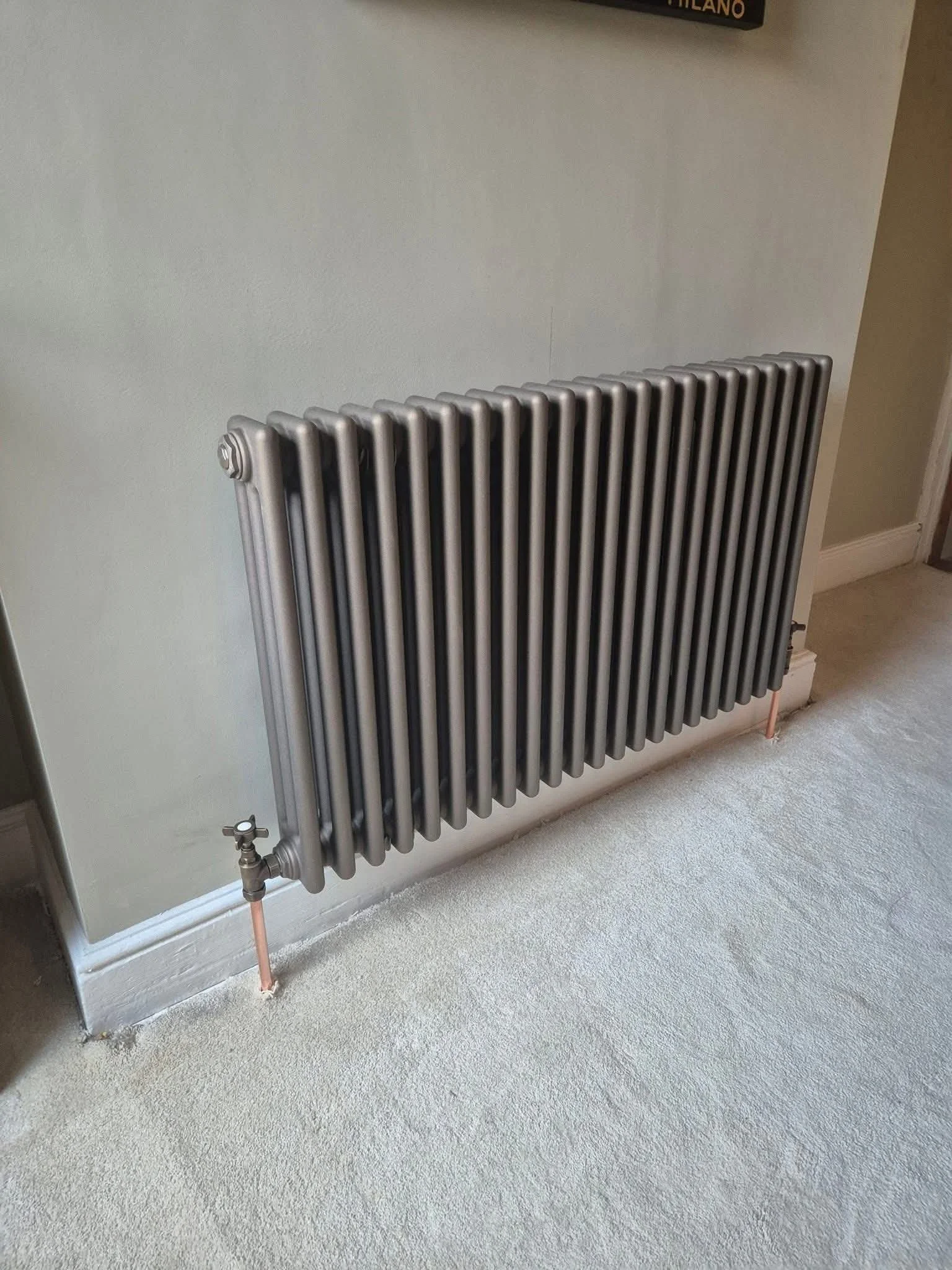 A metallic radiator mounted on a beige wall in a room with light-colored carpeted floor.