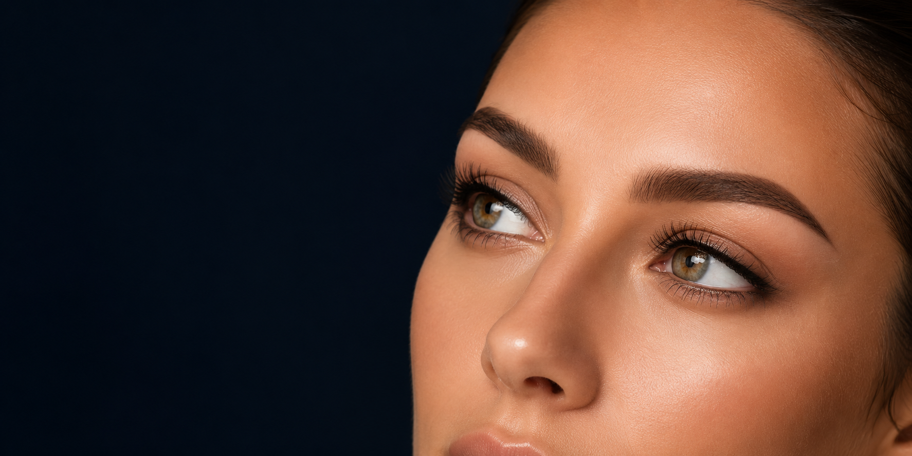 Close-up of a woman with softly defined ombré brows and natural lifted lashes against a deep navy background.