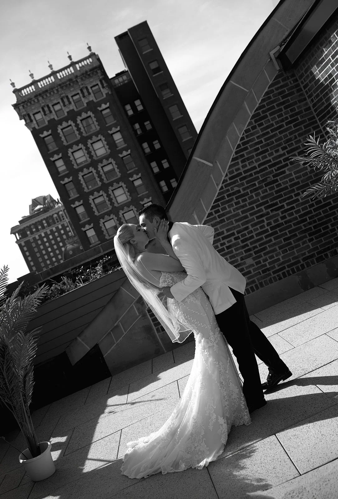 A black and white photo of a bride and groom kissing on a rooftop with city buildings in the background, leaning against a brick wall.