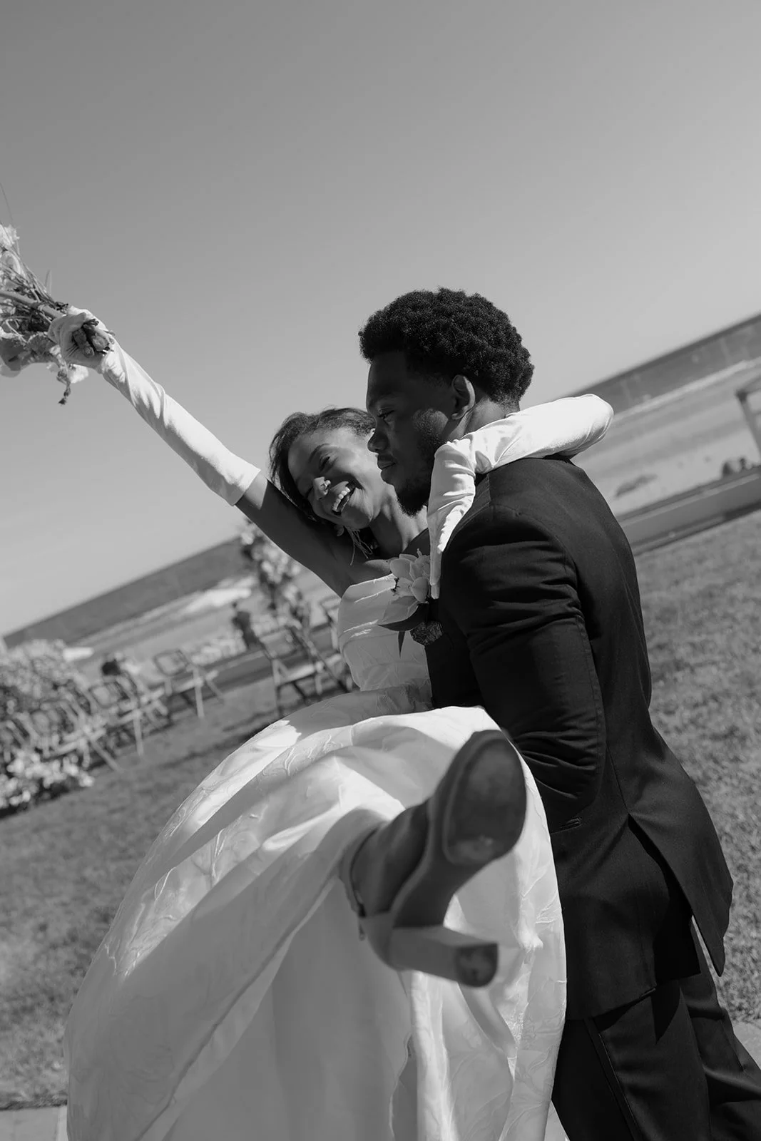A black-and-white photo of a couple kissing on a dock, dressed in formal attire, with boats and a marina in the background.