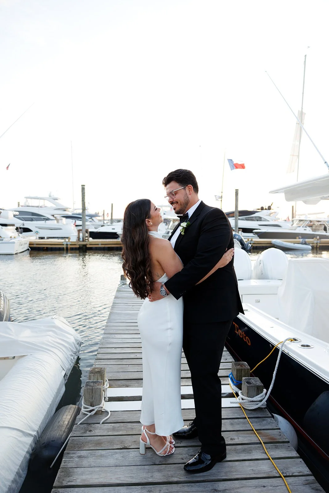 A newlywed couple dressed in wedding attire, embracing on a wooden dock at a marina with yachts and boats in the background.