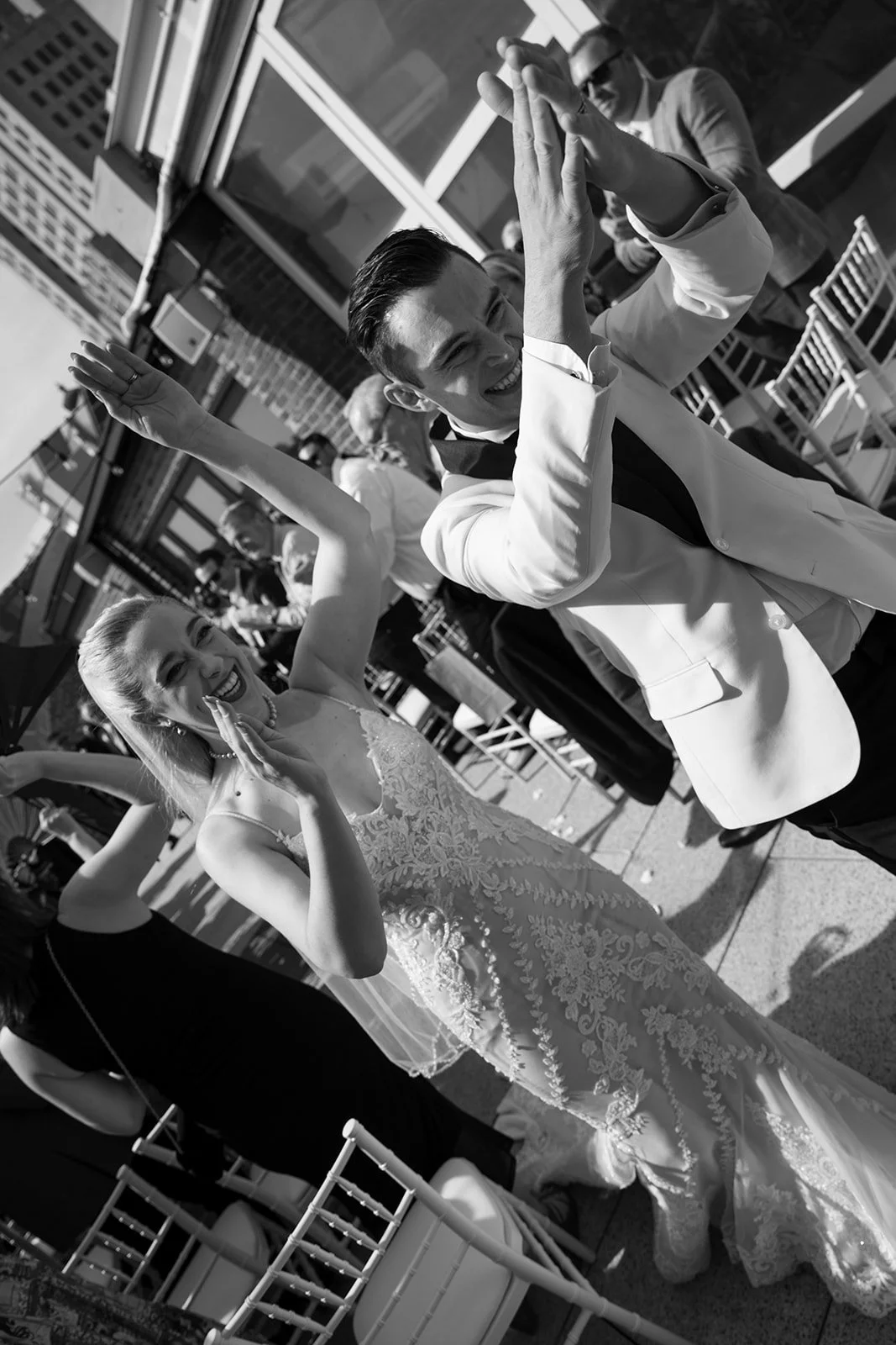 Black and white photo of a happy couple celebrating at a wedding reception outdoors, with the man in a tuxedo and bow tie and the woman in a lace wedding gown, both smiling and dancing.