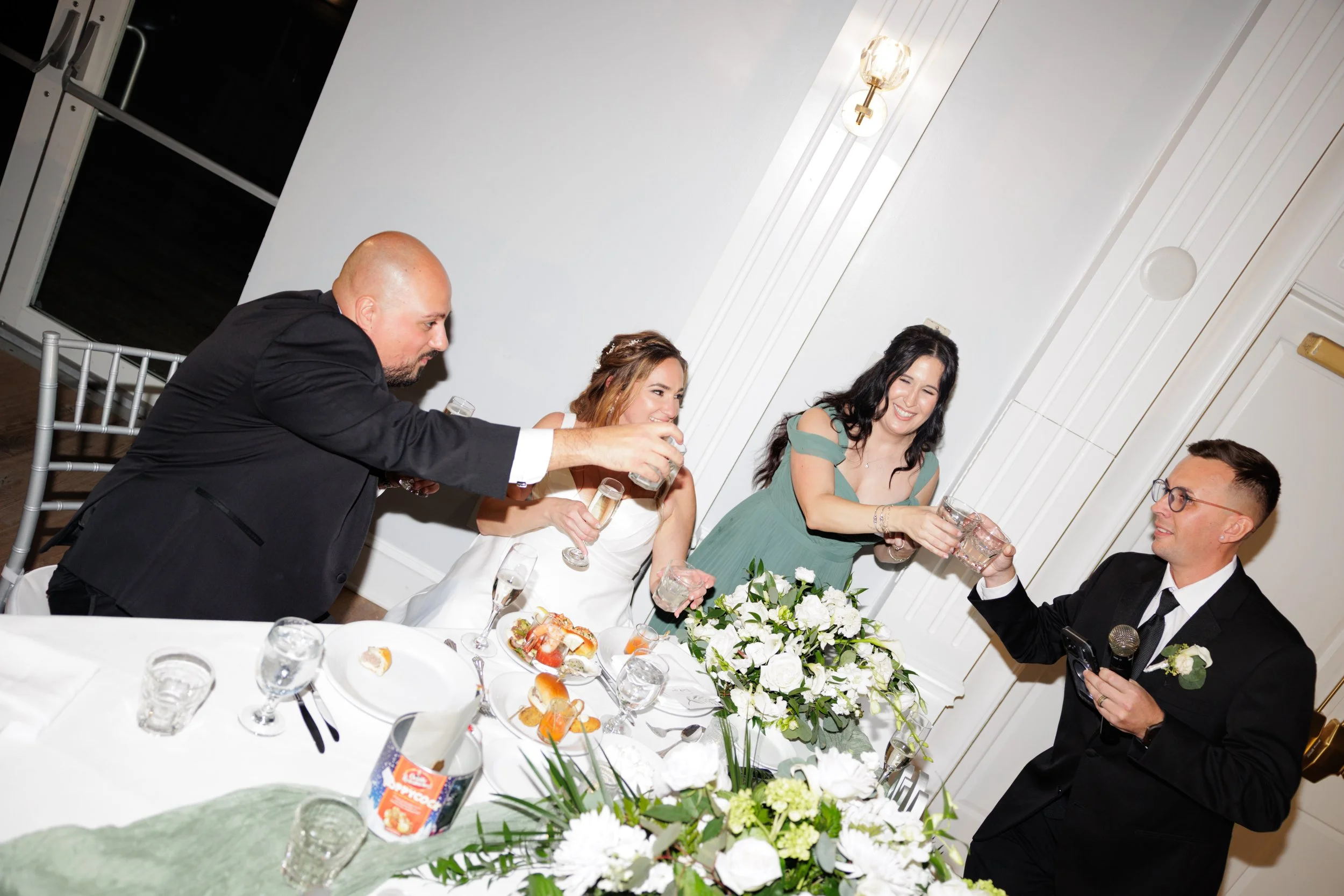 People gathered around a dinner table, toasting with glasses at a celebration event, with floral centerpiece and food on the table.