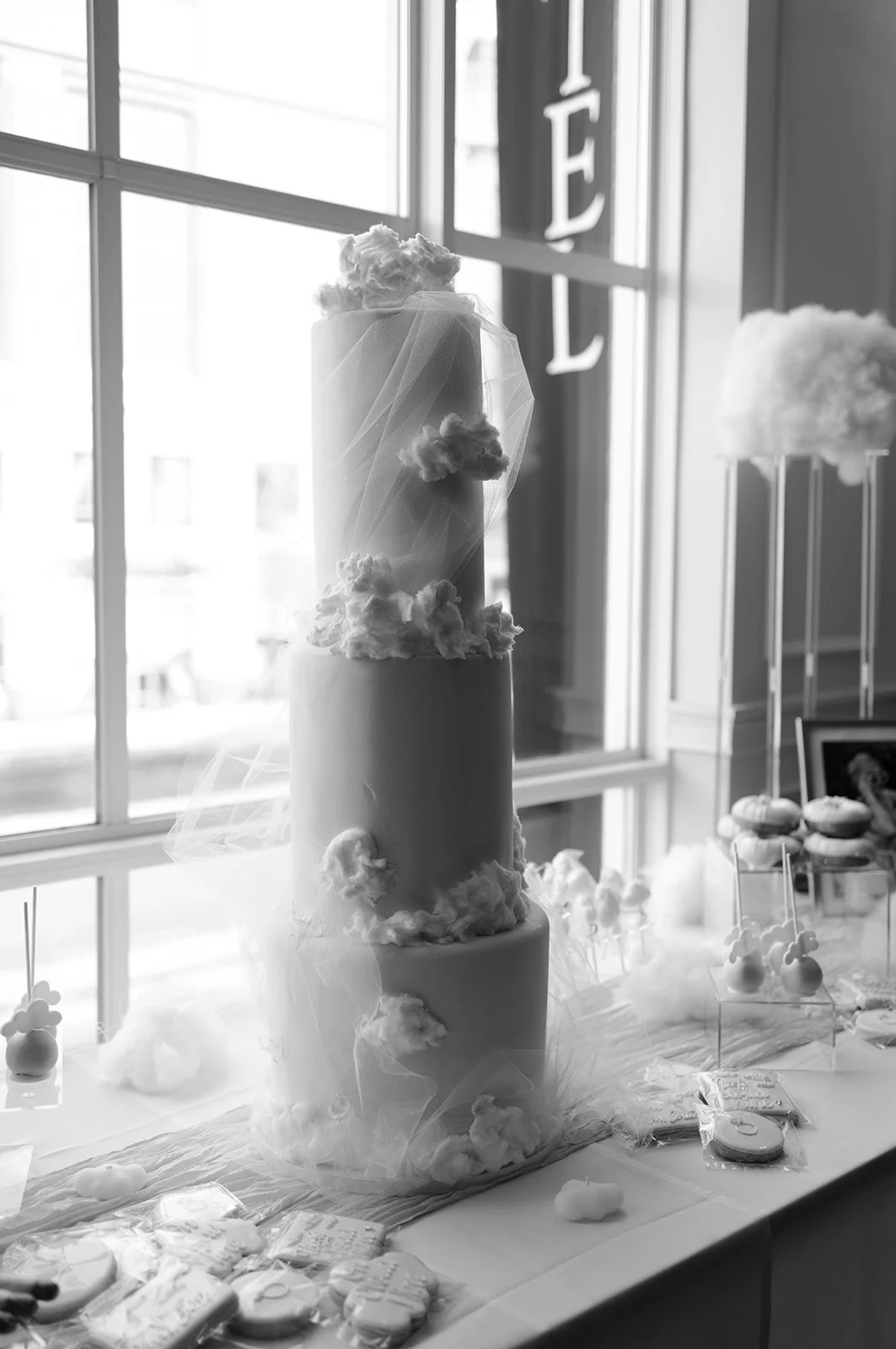 A four-tier wedding cake decorated with clouds and tulle, placed on a table decorated with cloud-shaped treats, with large windows and the word 'FEEL' hanging in the background.