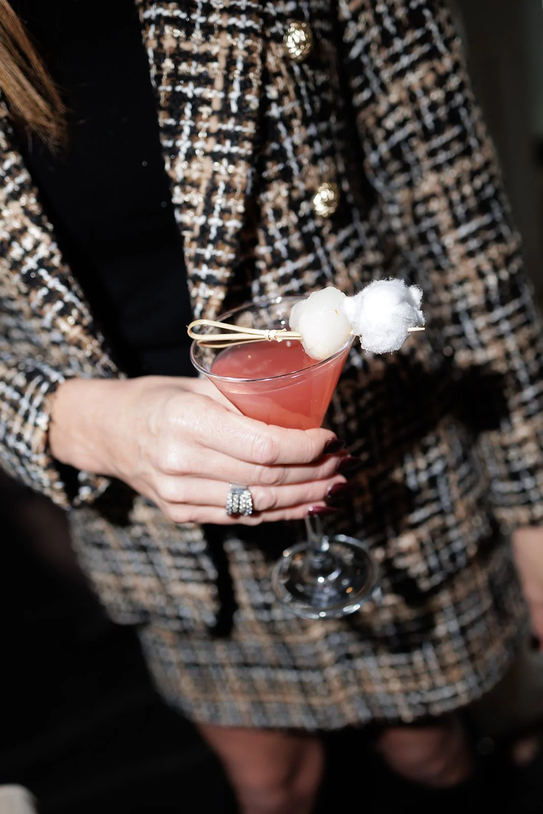 A woman wearing a black and gold tweed jacket is holding a pink cocktail in a martini glass garnished with cotton candy and a white ball, with her fingers showing a sparkling wedding ring.