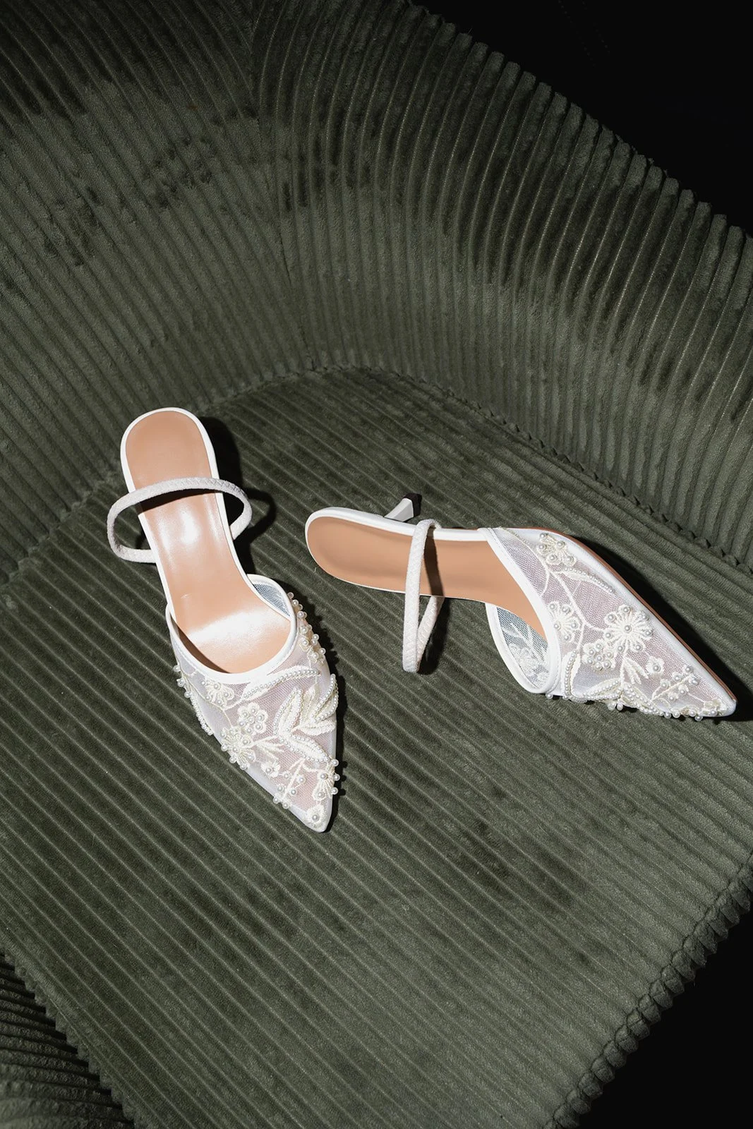 Pair of white embroidered high-heeled shoes with pointed toes and ankle straps on a gray textured chair.