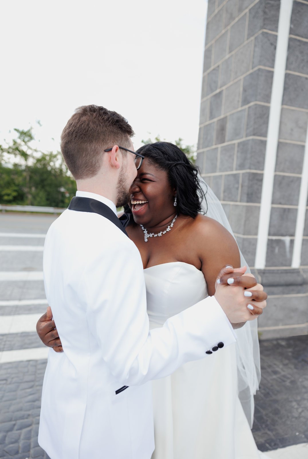 A newlywed couple sharing a joyful moment outdoors, with the bride wearing a strapless wedding gown and necklace, and the groom in a tuxedo, holding hands and touching foreheads.
