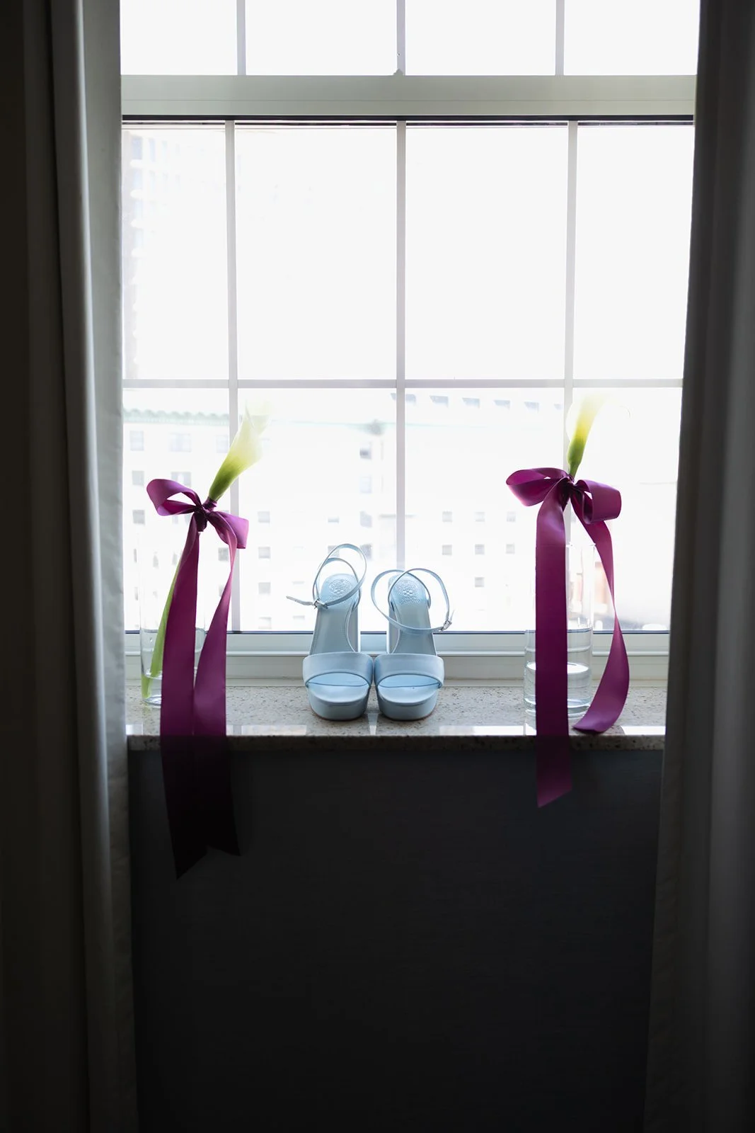 White high-heeled shoes with ankle straps placed on a windowsill between two tall vases decorated with pink ribbons and white calla lilies, all in front of a large window.