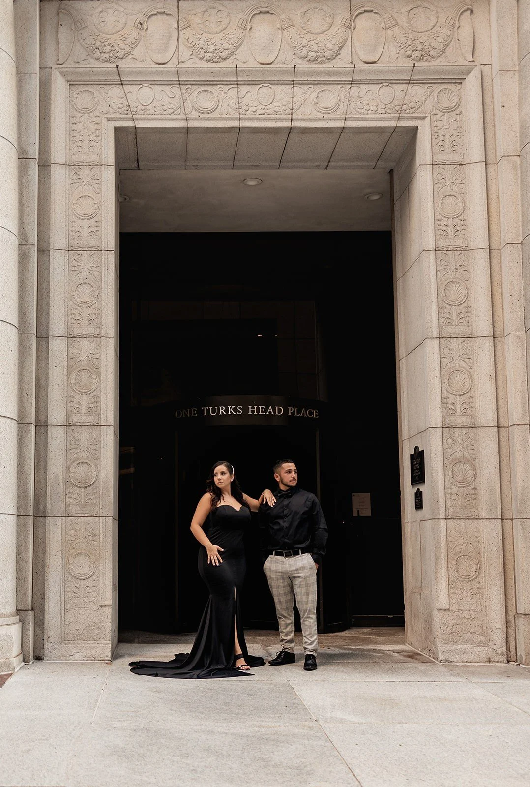 A man and woman dressed formally standing in front of the entrance to One Turks Head Place, a building with a grand stone archway with ornate carvings. The woman is wearing a long, black, strapless gown and the man is wearing a black shirt with plaid pants.