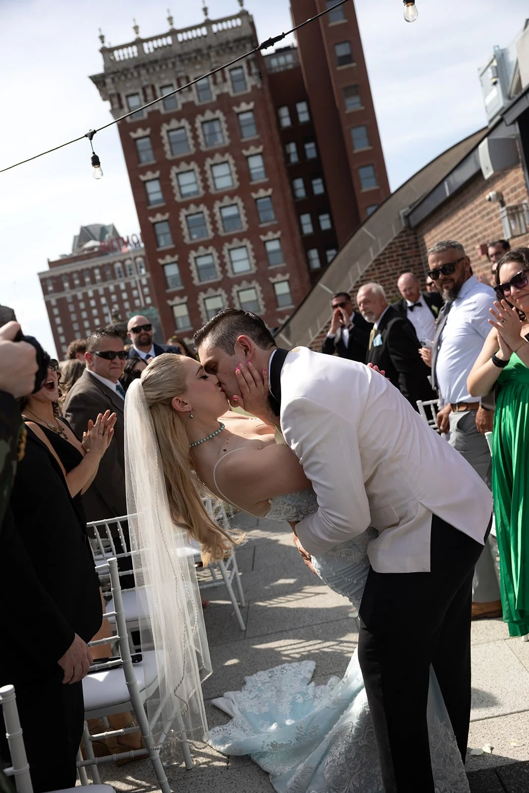 A bride and groom share a kiss during their wedding ceremony outdoors, surrounded by friends and family, with a cityscape featuring tall buildings in the background.