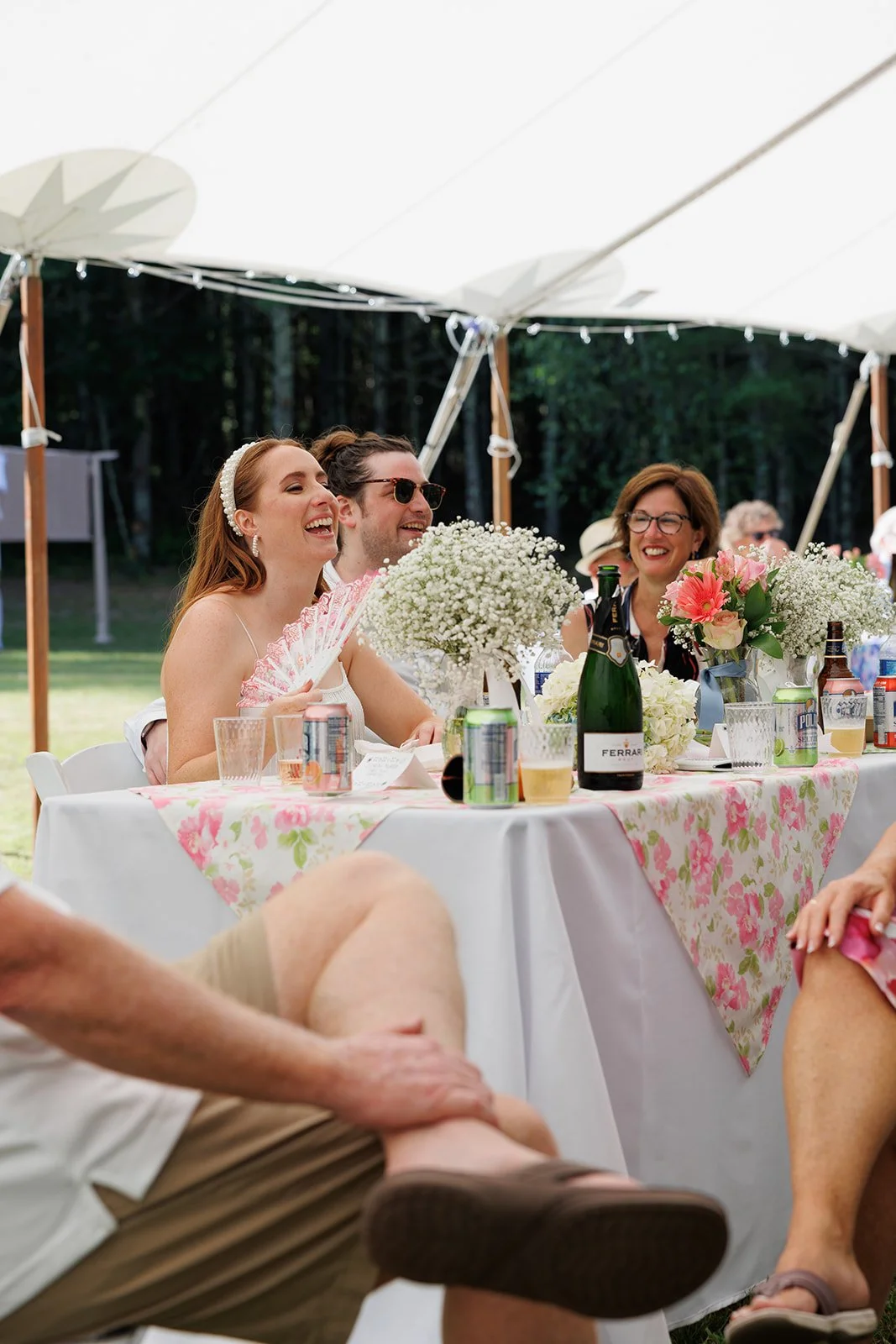 People enjoying a celebration outdoors under a white canopy, sitting at a decorated table with flowers, drinks, and food, laughing and socializing.