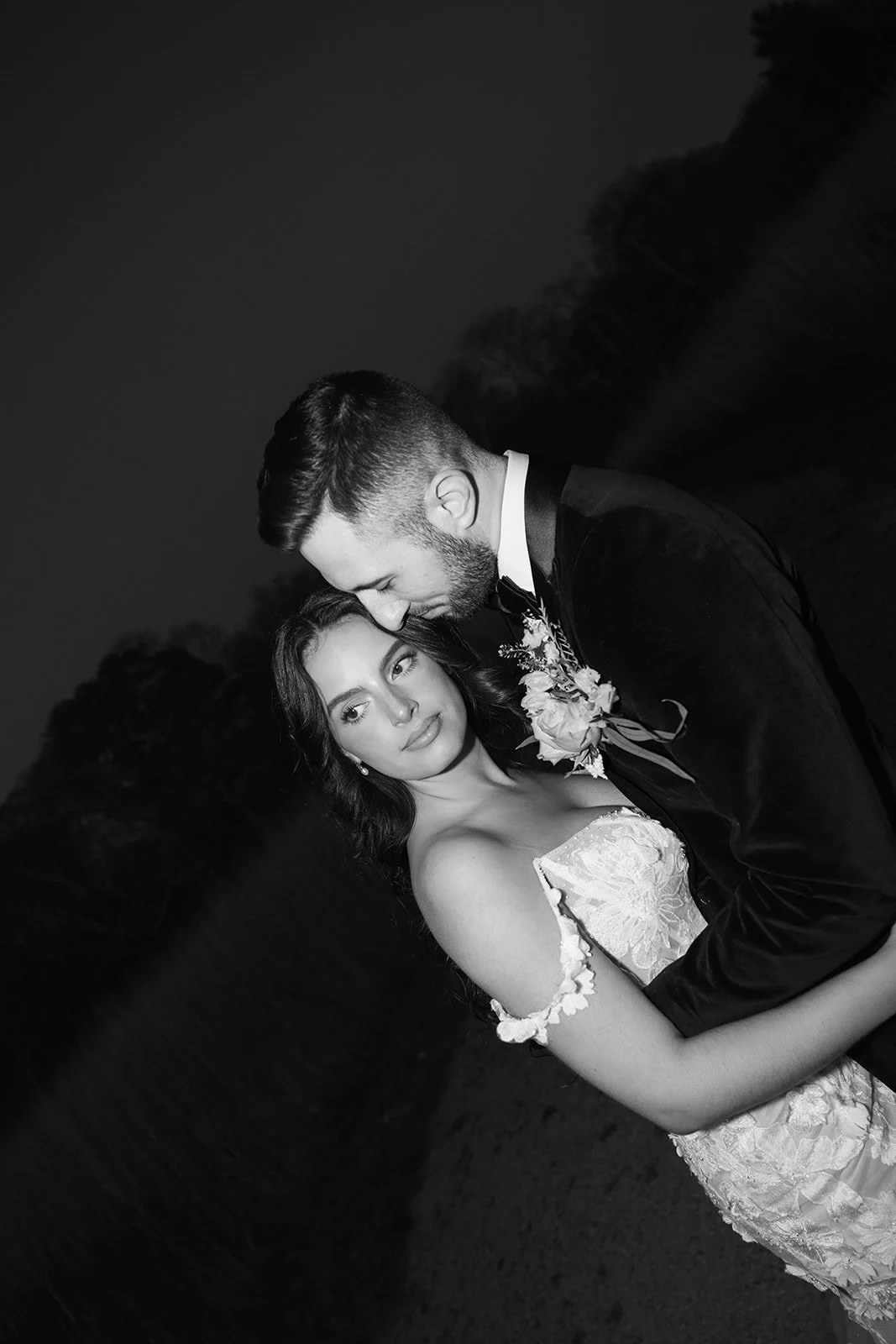 A black-and-white photo of a bride and groom embracing outdoors at night, with trees in the background. The groom is leaning in close with his forehead touching the bride's head, both smiling softly.