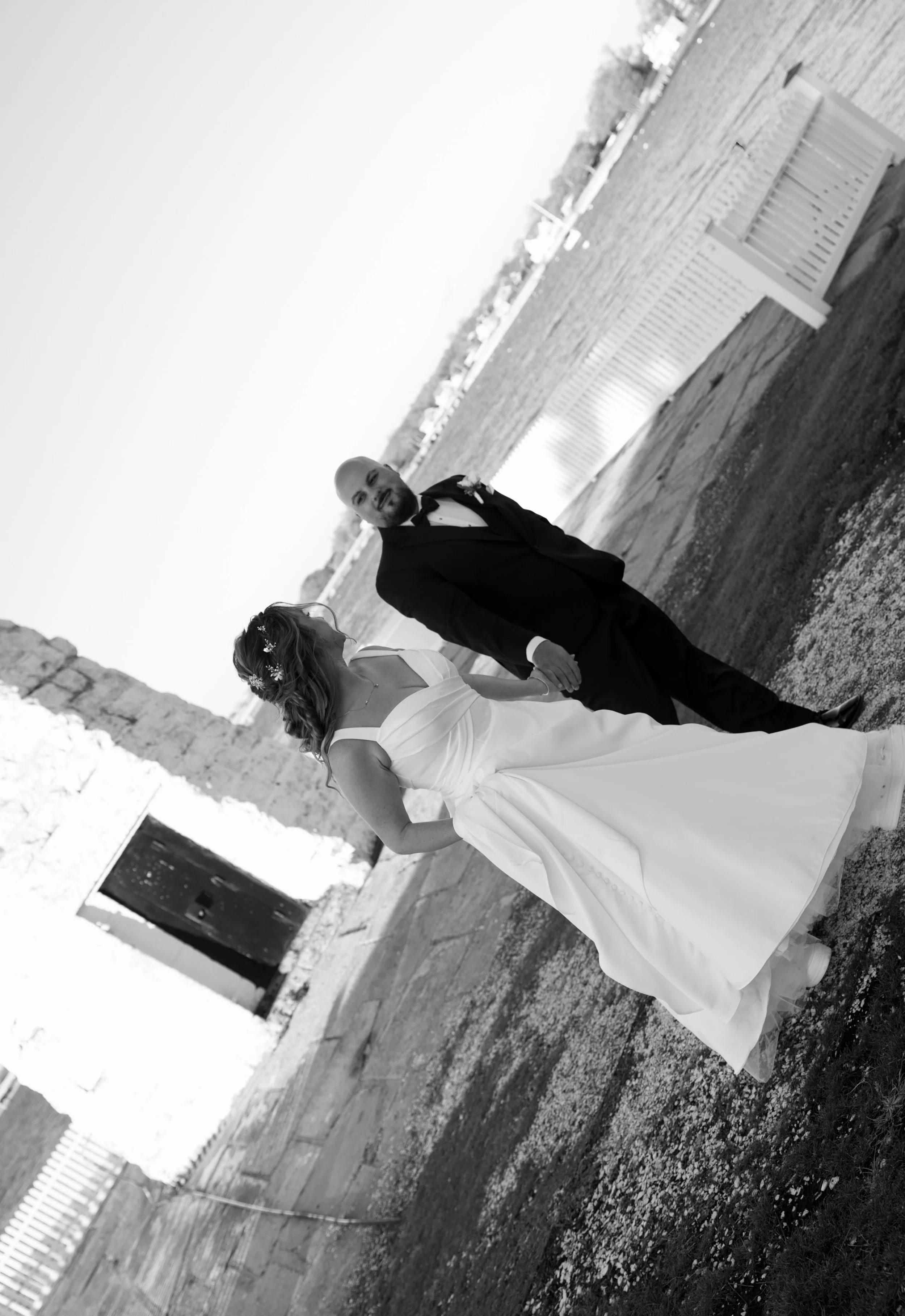A bride and groom holding hands outdoors, standing on a dirt path with a white fence on one side and a stone wall on the other. The bride is wearing a white wedding dress and a decorative hair accessory, and the groom is dressed in a dark suit with a white shirt and black bowtie. The sky is clear, and there is a distant view of trees and possibly a body of water.