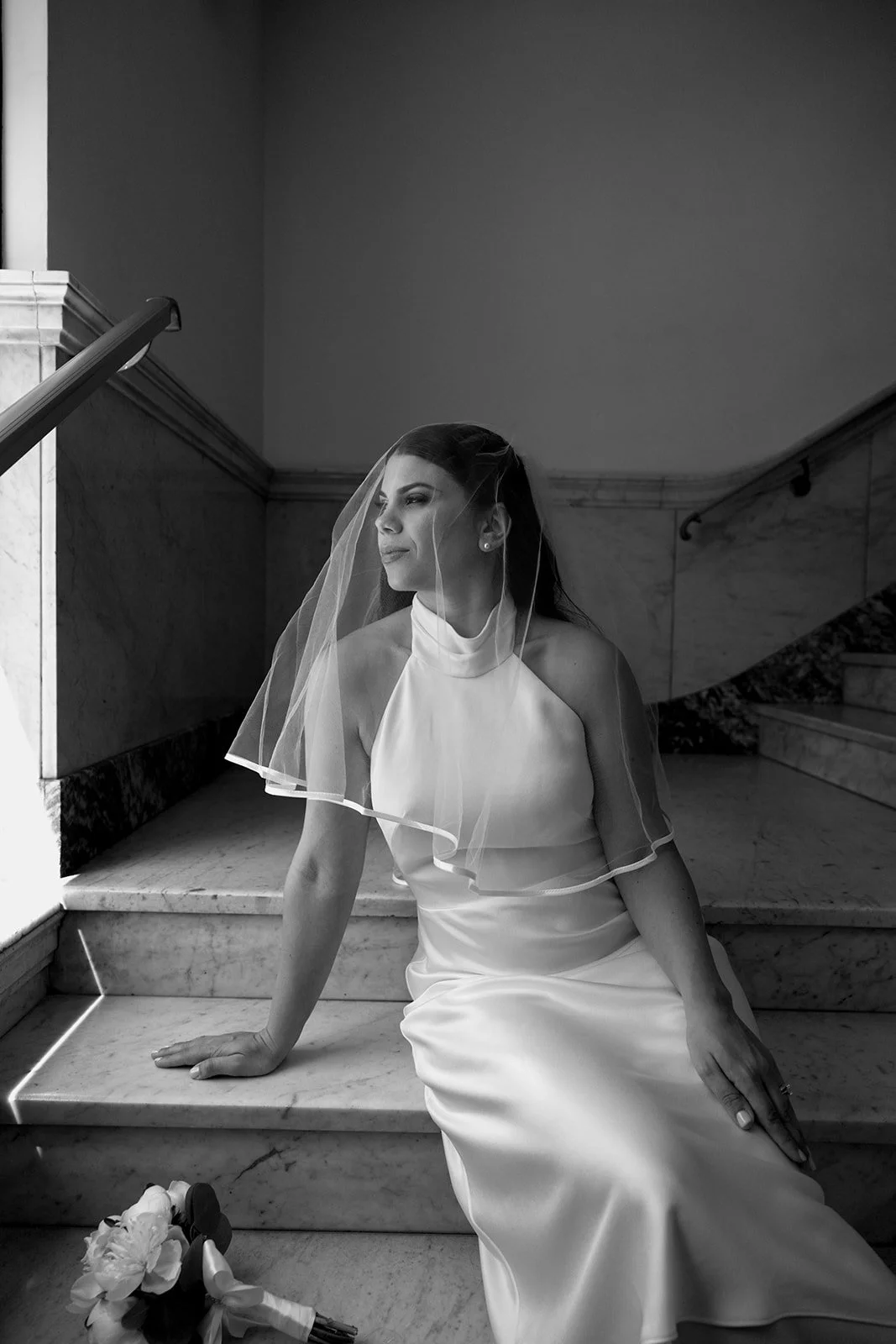 Black and white photo of a woman in a wedding dress and veil sitting on marble stairs, looking out. A bouquet is on the step nearby.