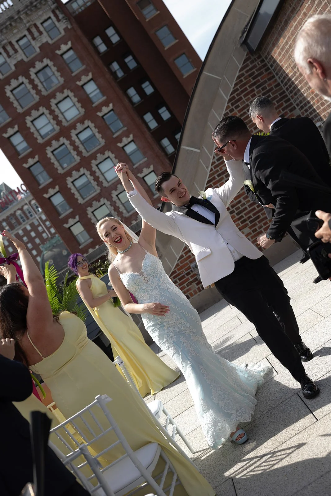 A group of wedding celebration attendees, including a bride in a white lace dress, a groom in a white tuxedo jacket, and bridesmaids in yellow dresses, dancing outdoors on a rooftop with city buildings in the background.
