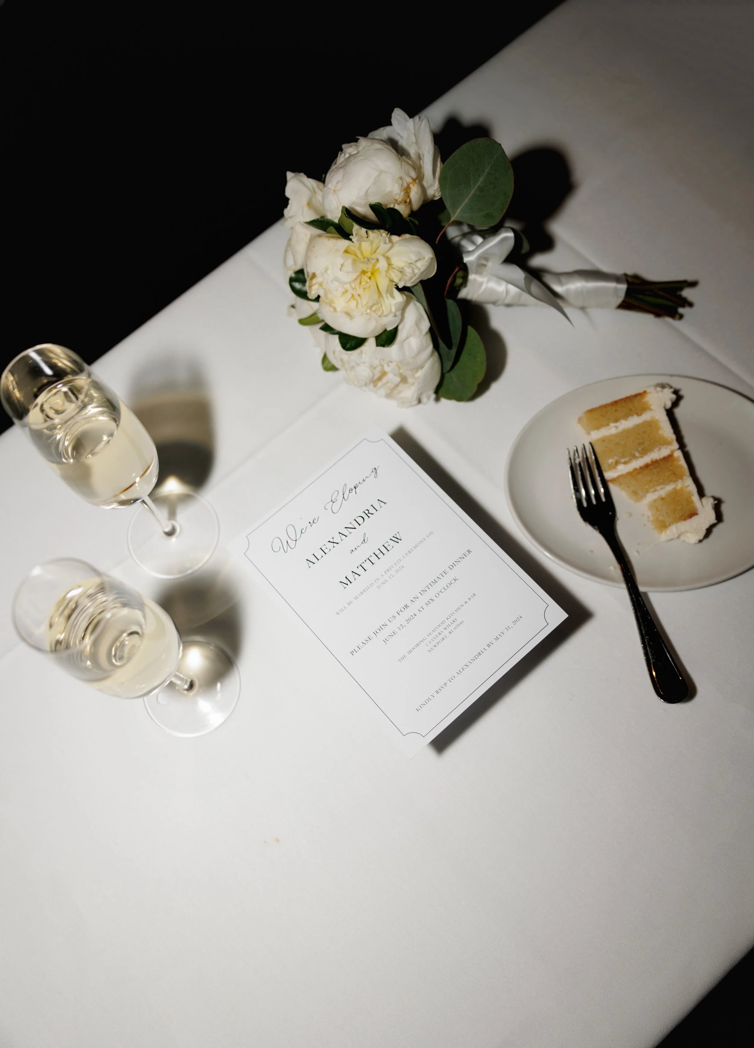 Wedding invitation on white table, floral boutonniere with white flowers and greenery, glass of champagne, and slice of cake on plate with fork, with a black and white background