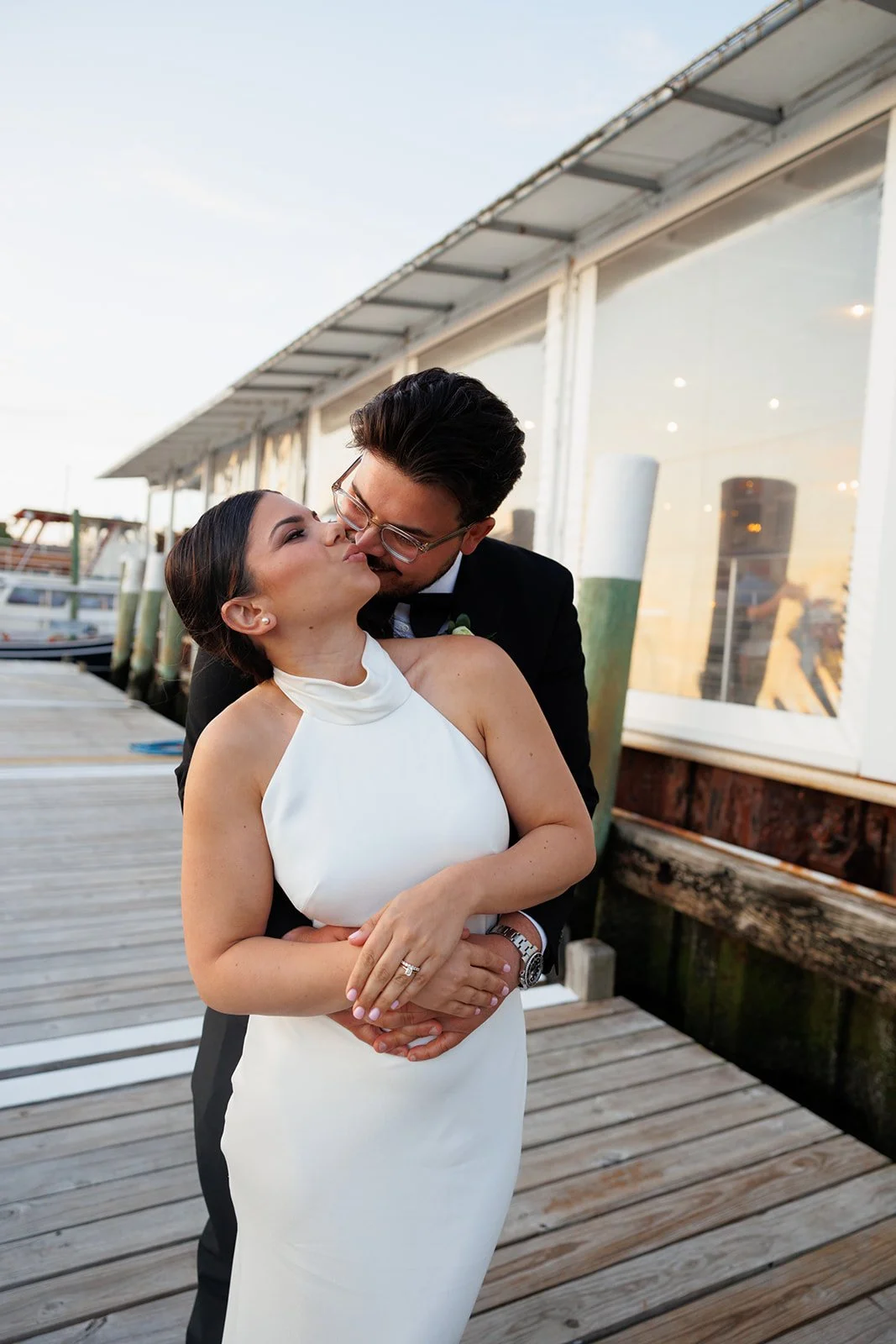 A couple dressed in wedding attire sharing a kiss on a wooden dock near water, with boats and a building in the background during sunset.