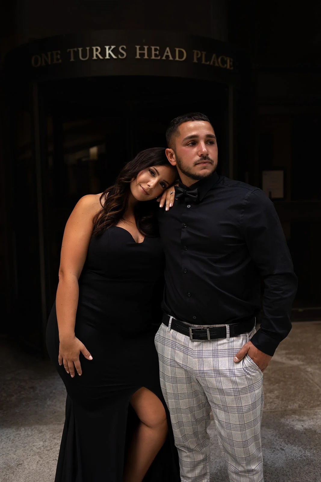 A woman leaning her head on a man's shoulder outside a building with a sign that reads 'One Turks Head Place.' The woman is wearing a black dress with a slit, and the man is wearing a black shirt, checkered pants, and a black bow tie.