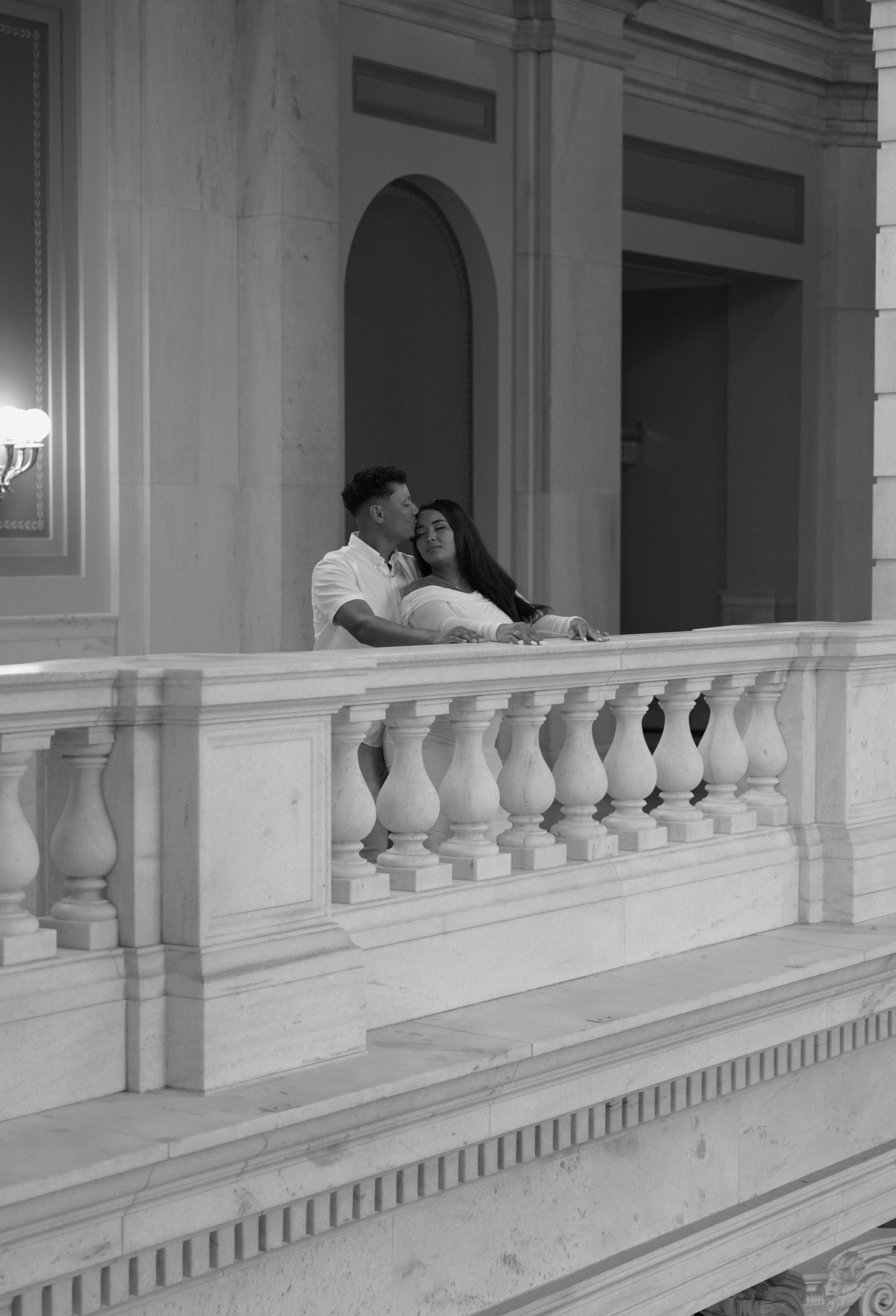 A black-and-white photograph of a man and a woman leaning on a marble balcony inside a grand building with ornate architectural features, including arches and columns.