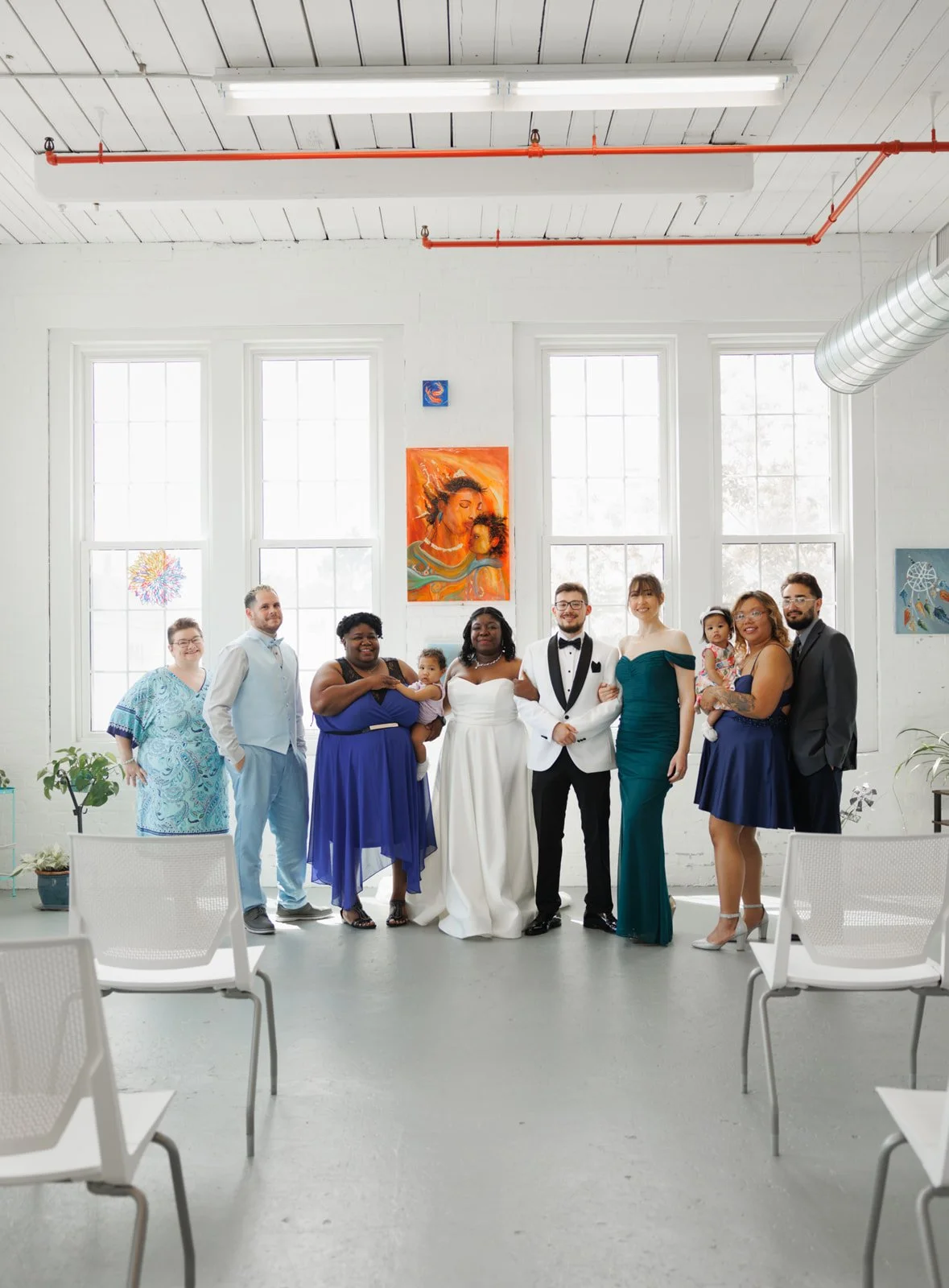 Group of eight people at a wedding reception, standing indoors in front of large windows with artwork on the walls, business casual and formal attire, several holding children.