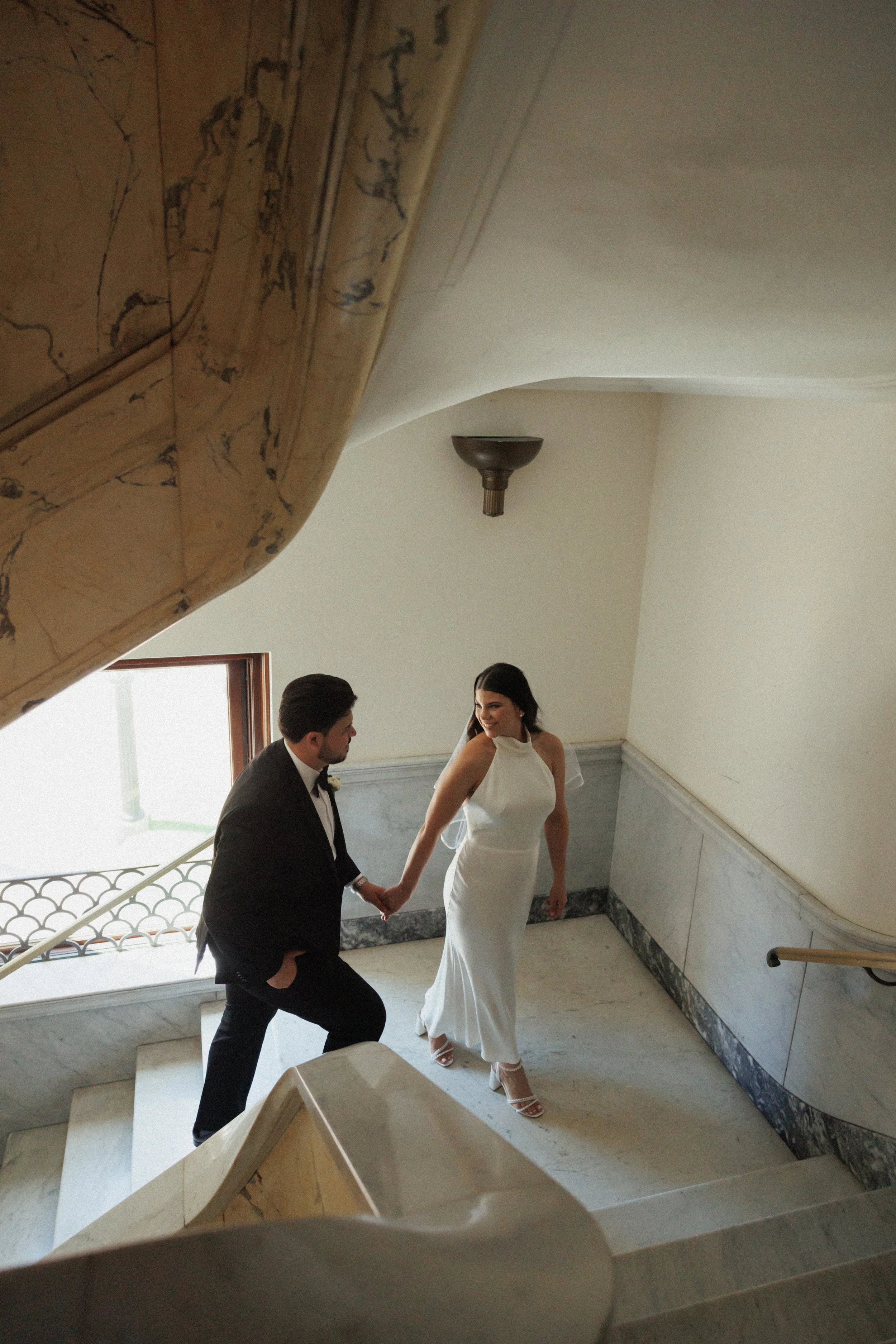 A man and woman holding hands on a staircase in an elegant interior, the woman in a white dress and the man in a black tuxedo. They are smiling and looking at each other.