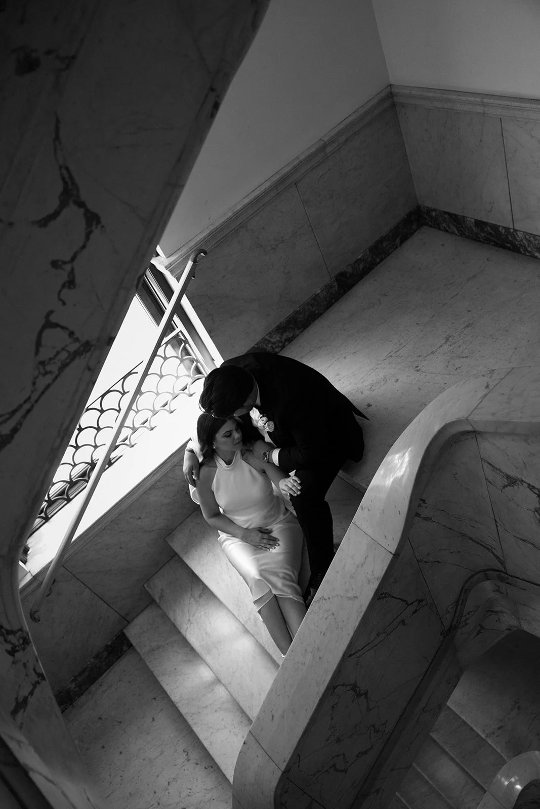 Black and white photo of a couple on a staircase, with the man leaning down to kiss the woman, who is sitting on the stairs, inside a marble-walled building near a window.