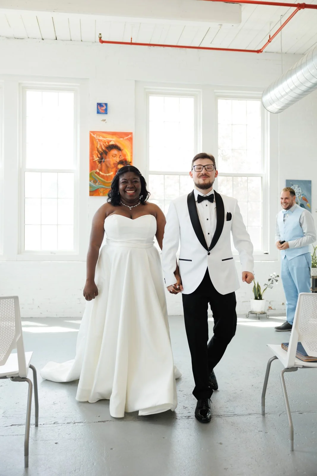 Couple dressed in wedding attire holding hands and smiling, with a man in a light blue suit in the background smiling, inside a bright room with white walls and large windows.