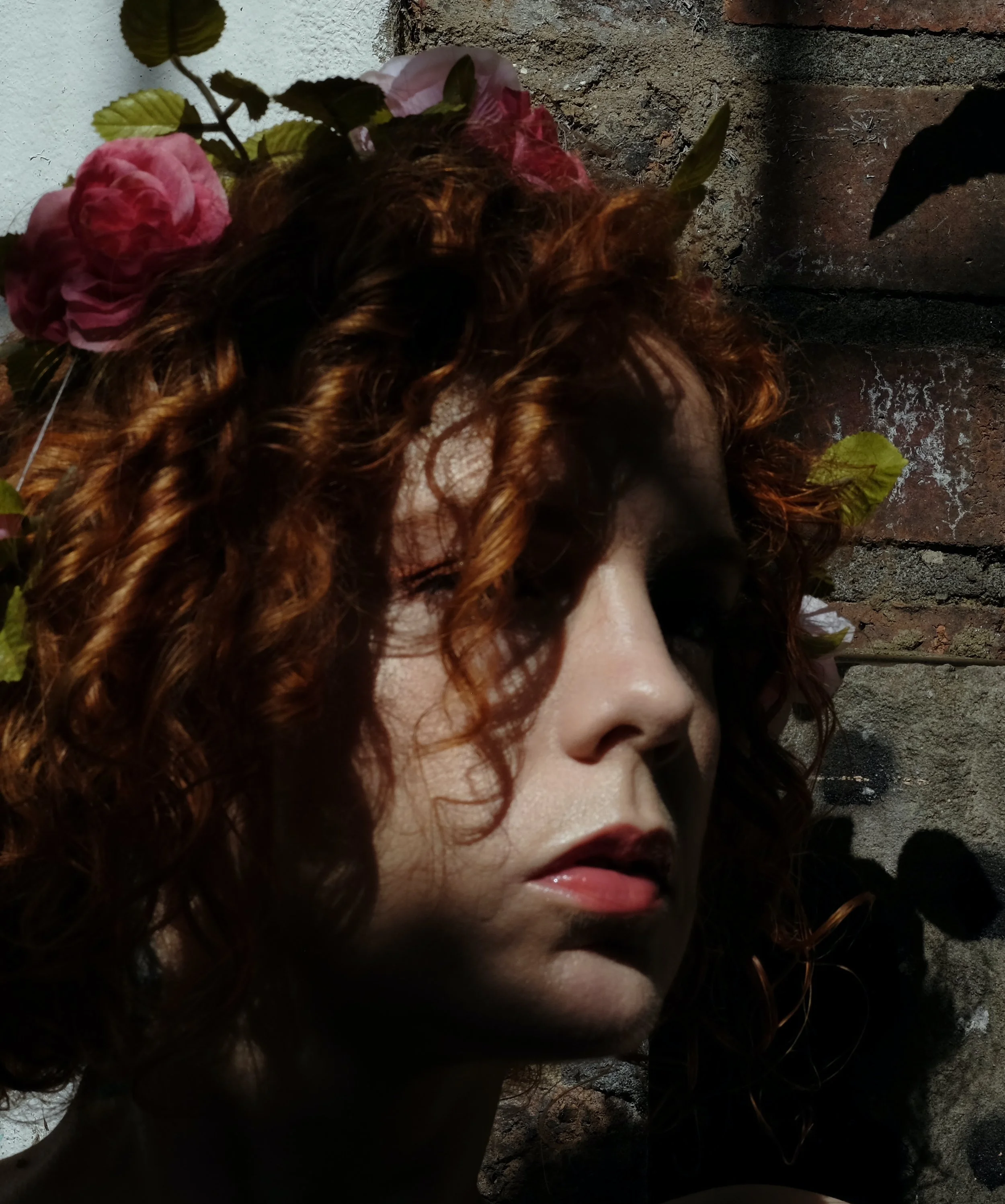 Close-up of a woman with curly red hair, adorned with pink flowers, lying against a stone wall.