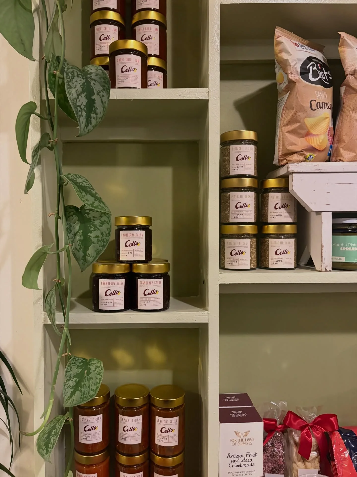 Shelf with jars labeled 'Cranberry Salsa' and 'Eggplant Relish.' House made Cello produce, all made in house, small batch using local ingredients. Hand poured into glass jars. Available for retail sale and can post