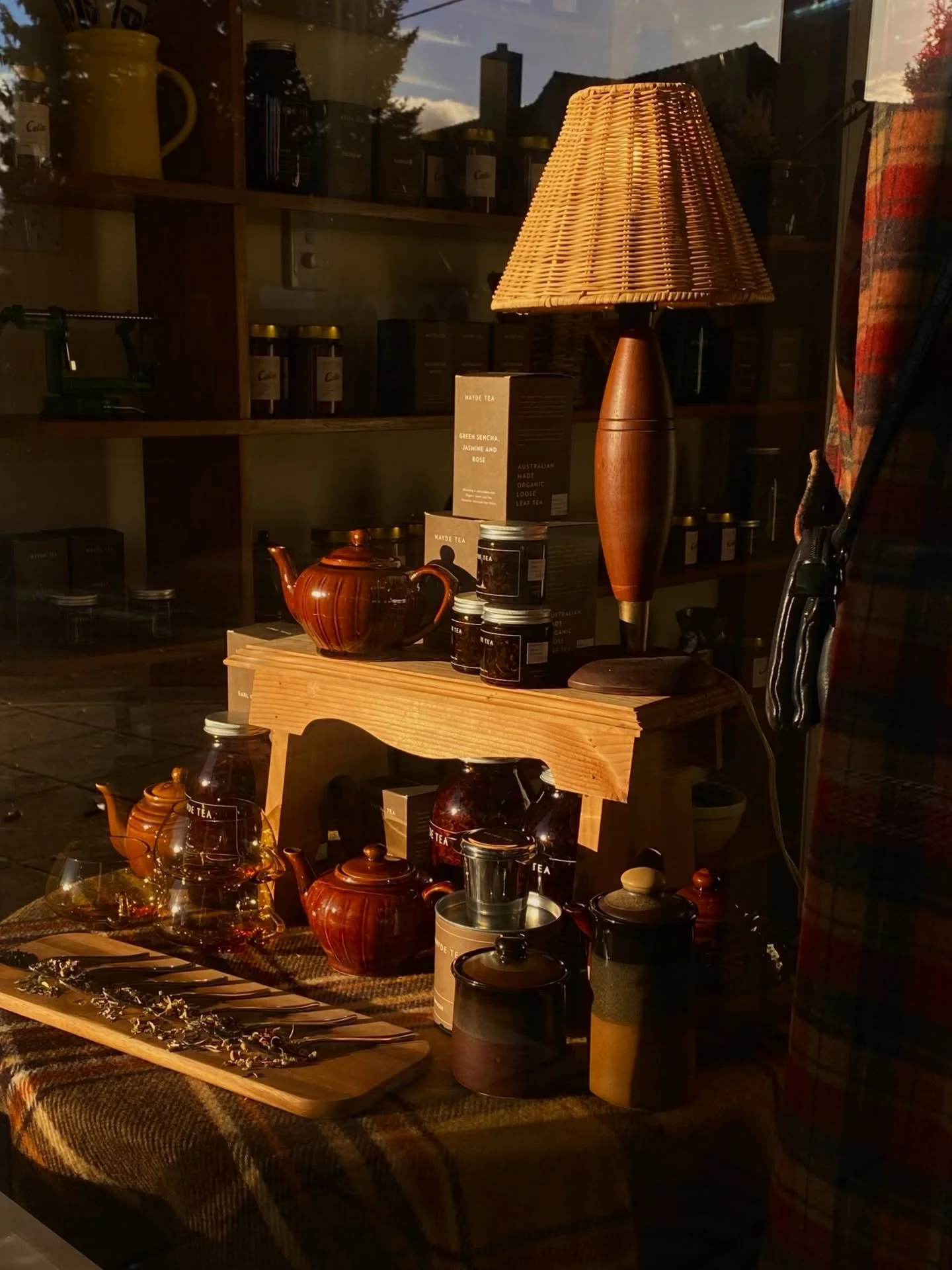 Display of boutique loose leaf tea collection by Mayde Tea. Set with vintage tea accessories including teapots, cups, jars, and herbal bundles in a store window with a wooden table and warm lighting.
