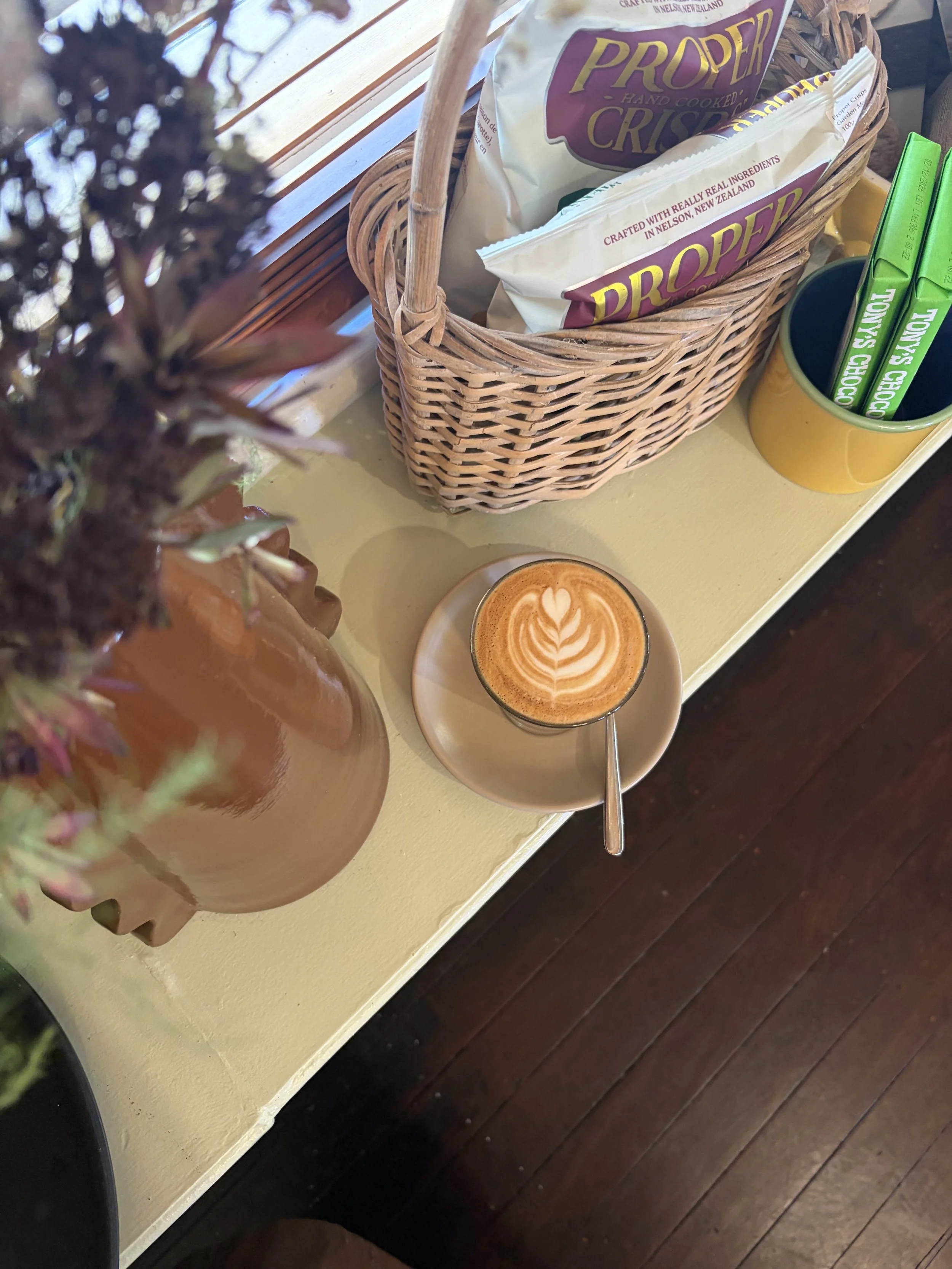 A cup of coffee with latte art on a saucer on a table, with a basket of snack packets and a cup holding green stirrers nearby.