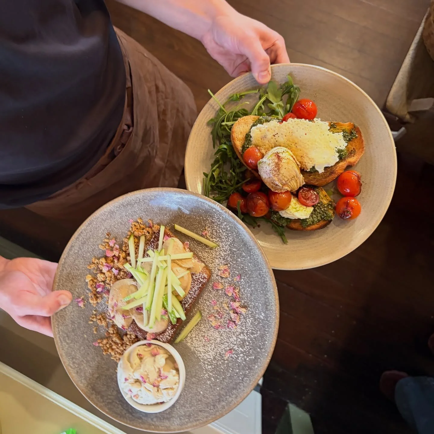 Two plates of gourmet food being presented. The first plate contains roasted vegetables, cherry tomatoes, and leafy greens. The second plate has a dessert topped with sliced fruit, nuts, and edible flowers, with a small side of cream.