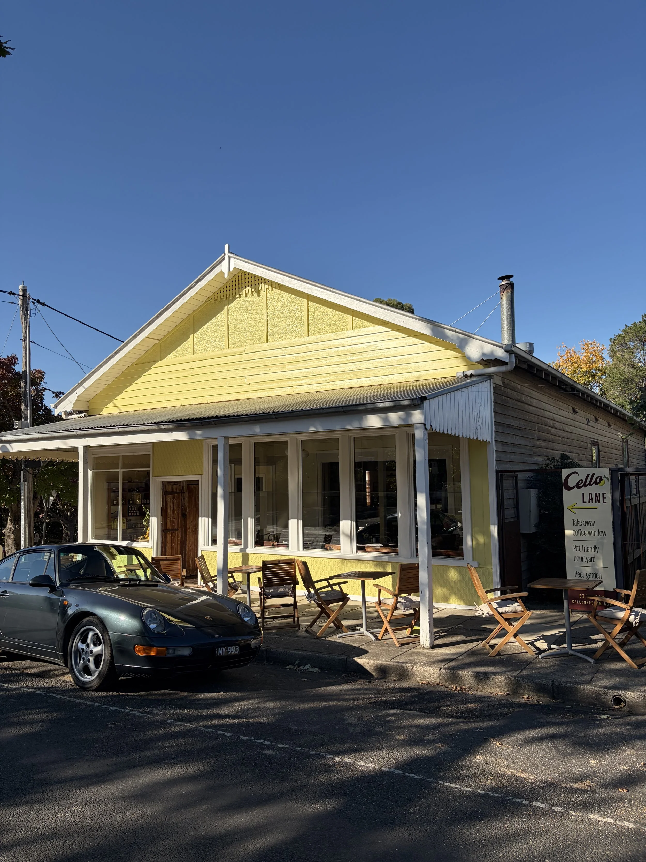 Exterior of a yellow building with large glass windows, outdoor tables and chairs, parked black sports car, and a sign pointing to a lane with take away coffee, pet friendly courtyard, beer garden, and other amenities. Clear blue sky, trees with some