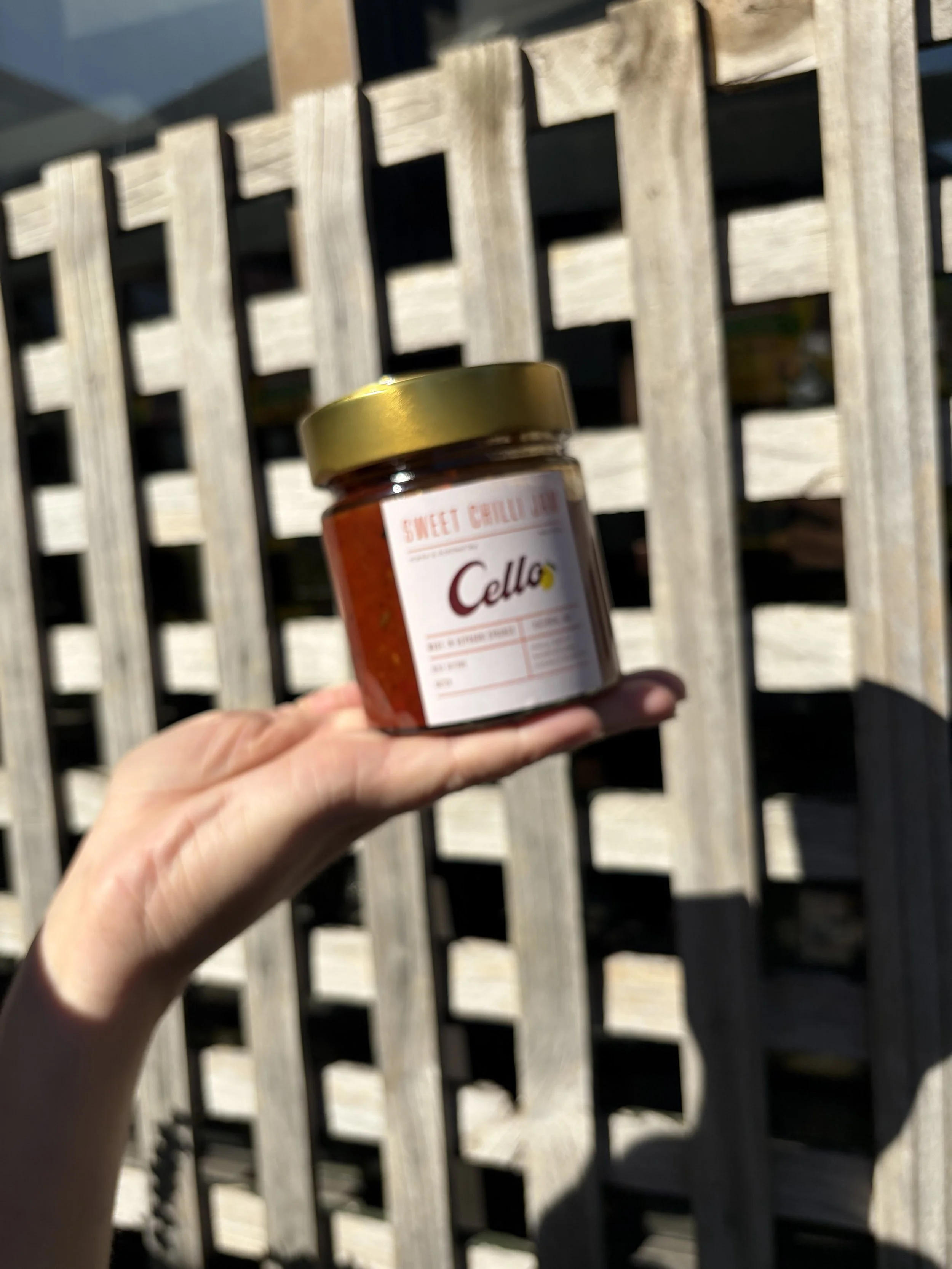 Close-up of a hand holding a jar of Cello's house made produce collection, Chilli Jam in front of a wooden fence, with shadows cast by sunlight.