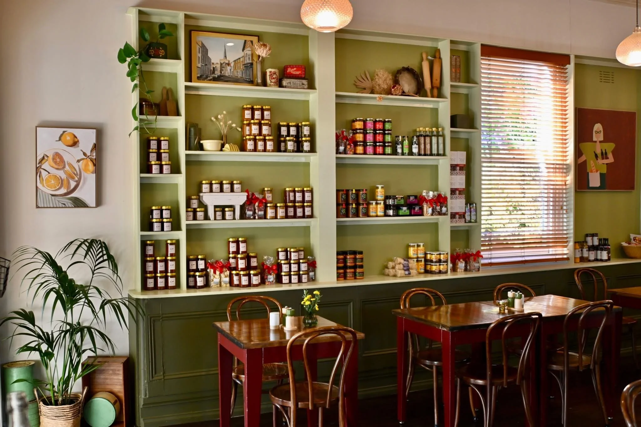 Interior of a cozy café with a large green shelf filled gourmet local foods and imported foods amongst vintage homewares. Retail shelving lining the walls of the cafe with vintage style venetain blinds. Classic Bentwood chairs and timber tables