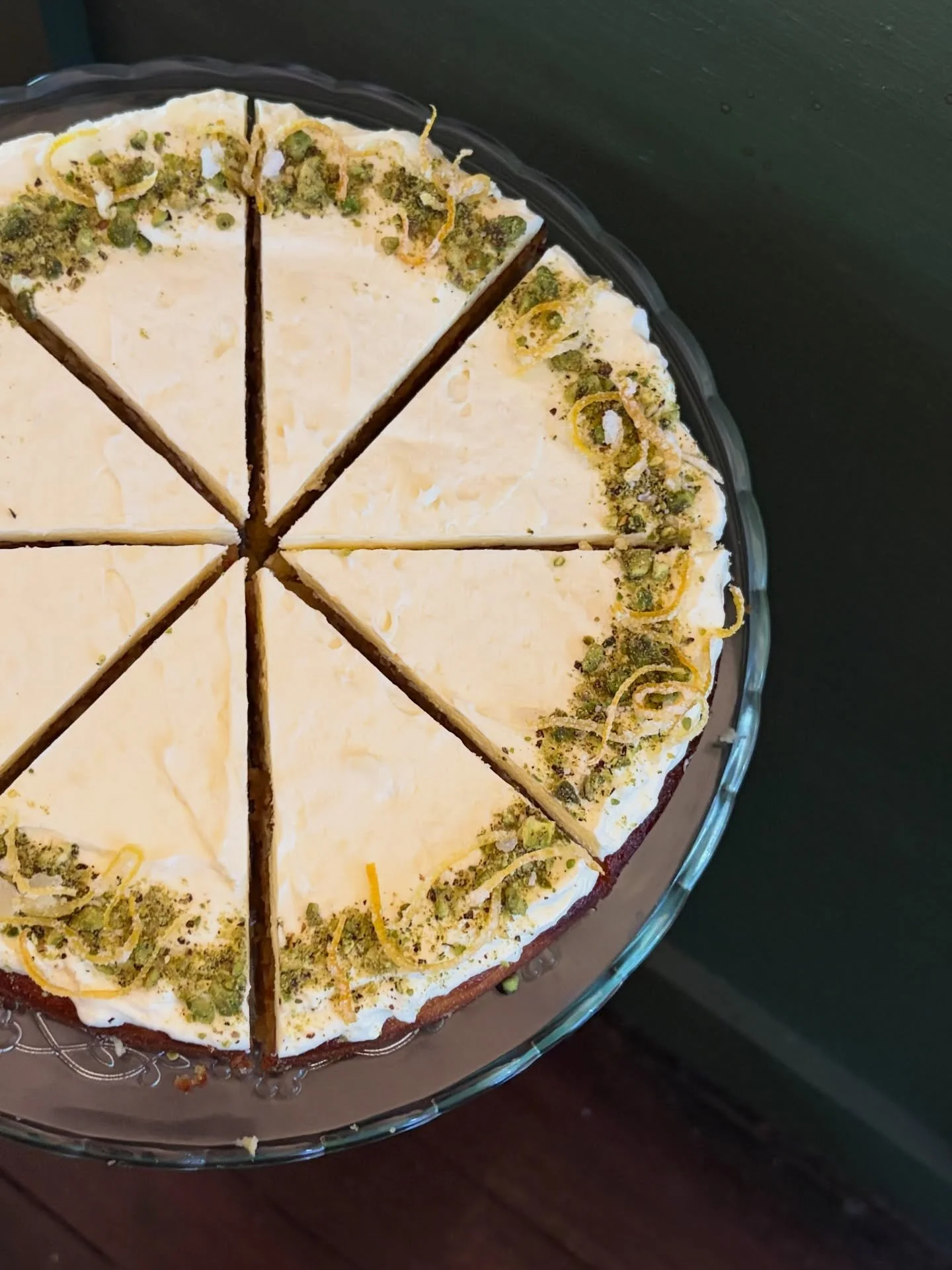 A round flourless orange cake topped with cream cheese frosting and garnished with chopped pistachios and lemon zest, sliced into six pieces. This cake is gluten free