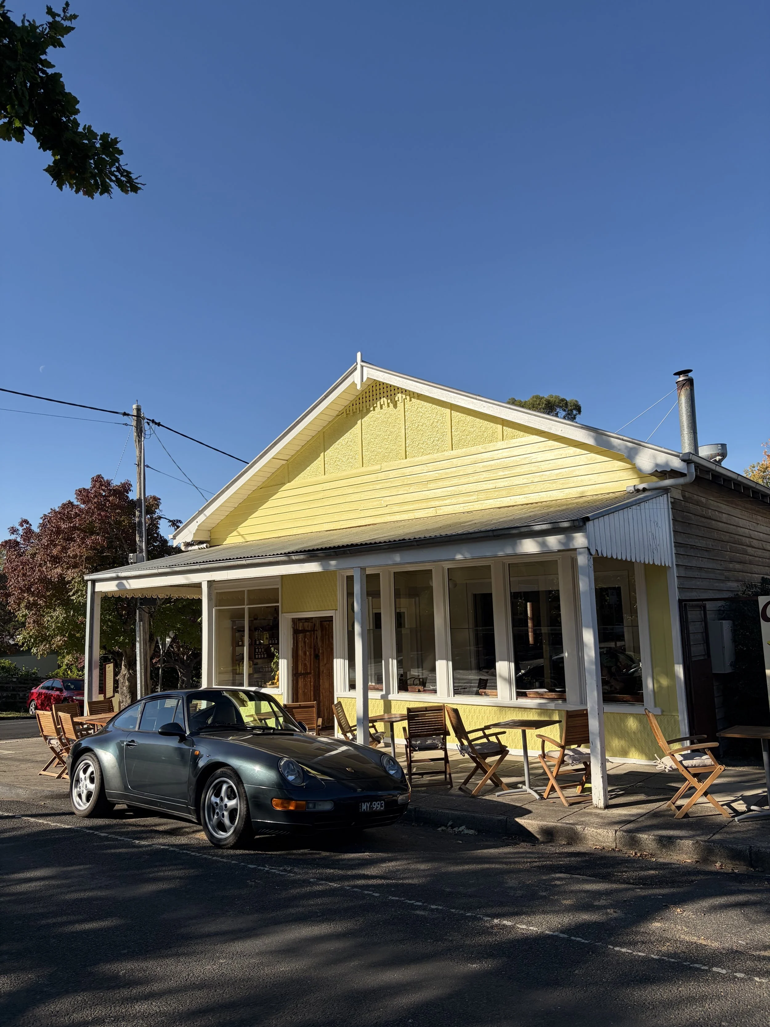 A yellow heritage weatherboard building with a white verandah and large windows, with outdoor dining seating and a vintage porche sports car parked in front, under a clear blue sky.