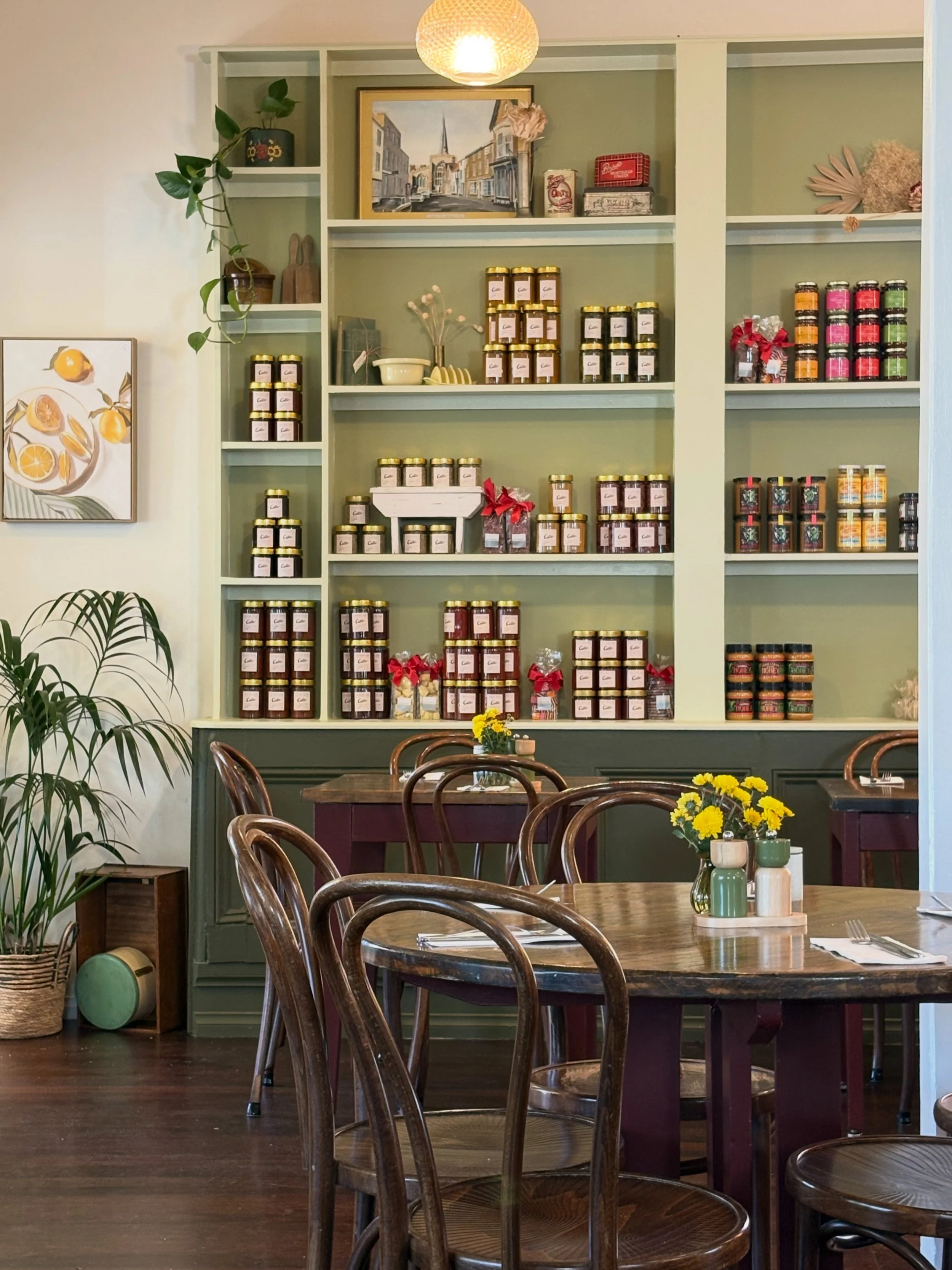 Interior of a cozy cafe or restaurant with wooden tables and chairs, a green shelving unit against the wall filled with jars and decor, potted plants, and a framed lemon-themed artwork on the wall.