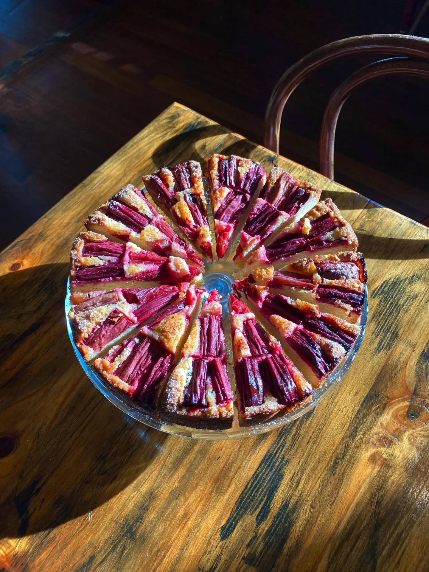 A round rhubarb frangipane tart sliced into individual pieces on a glass cake stand, placed on a wooden table with natural light.