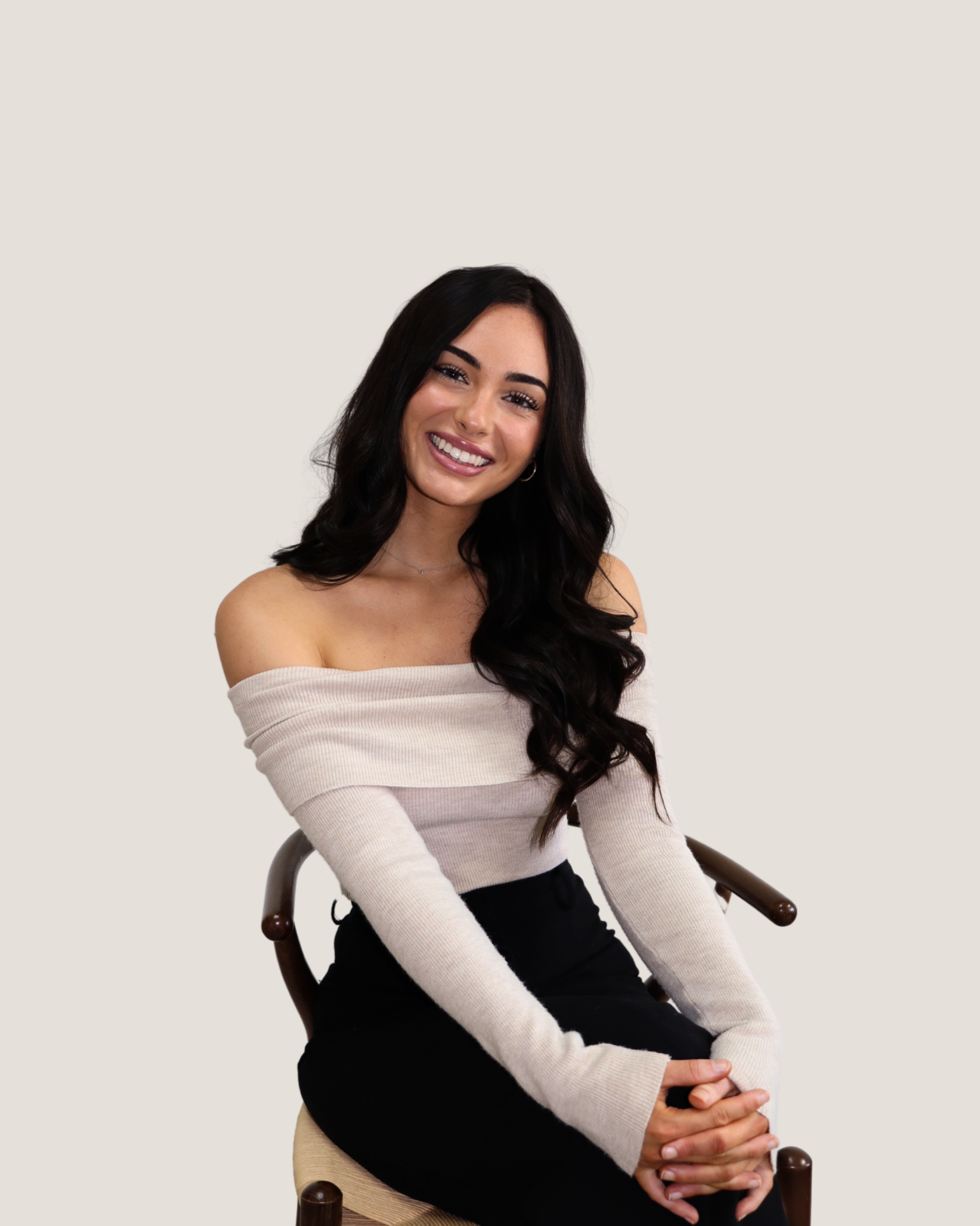 A woman with long dark hair, wearing a beige off-the-shoulder sweater and black pants, sitting on a wooden chair against a plain light background, smiling at the camera.