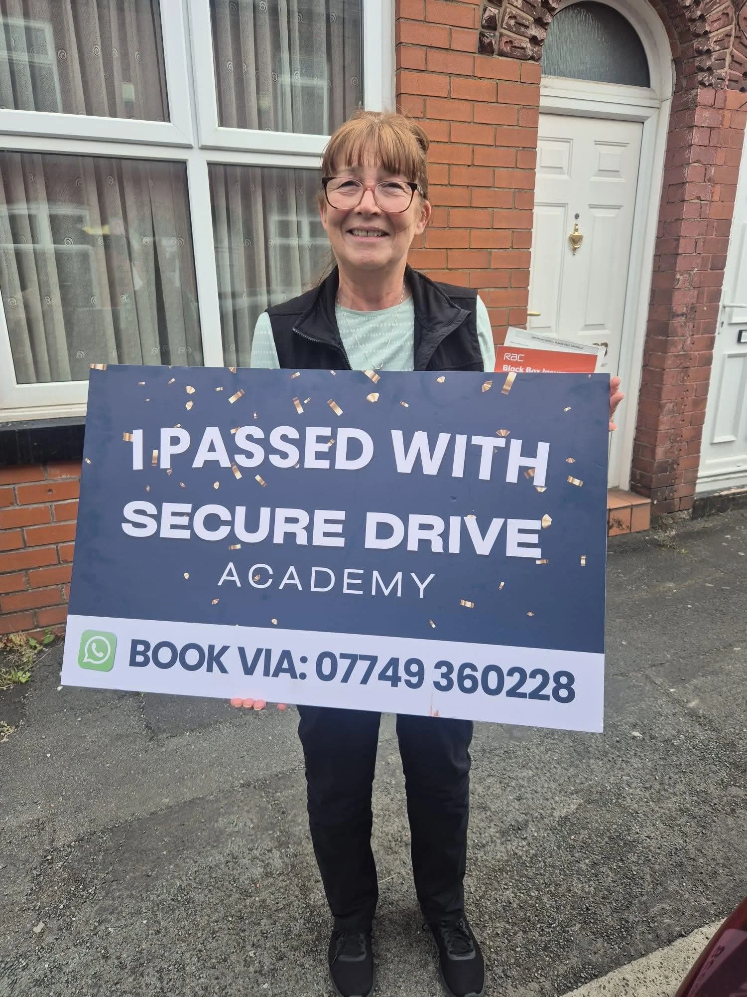 A woman standing outside in front of a brick house, holding a sign that reads 'I PASSED WITH SECURE DRIVE ACADEMY' and contact instructions, smiling and wearing glasses and casual clothing.