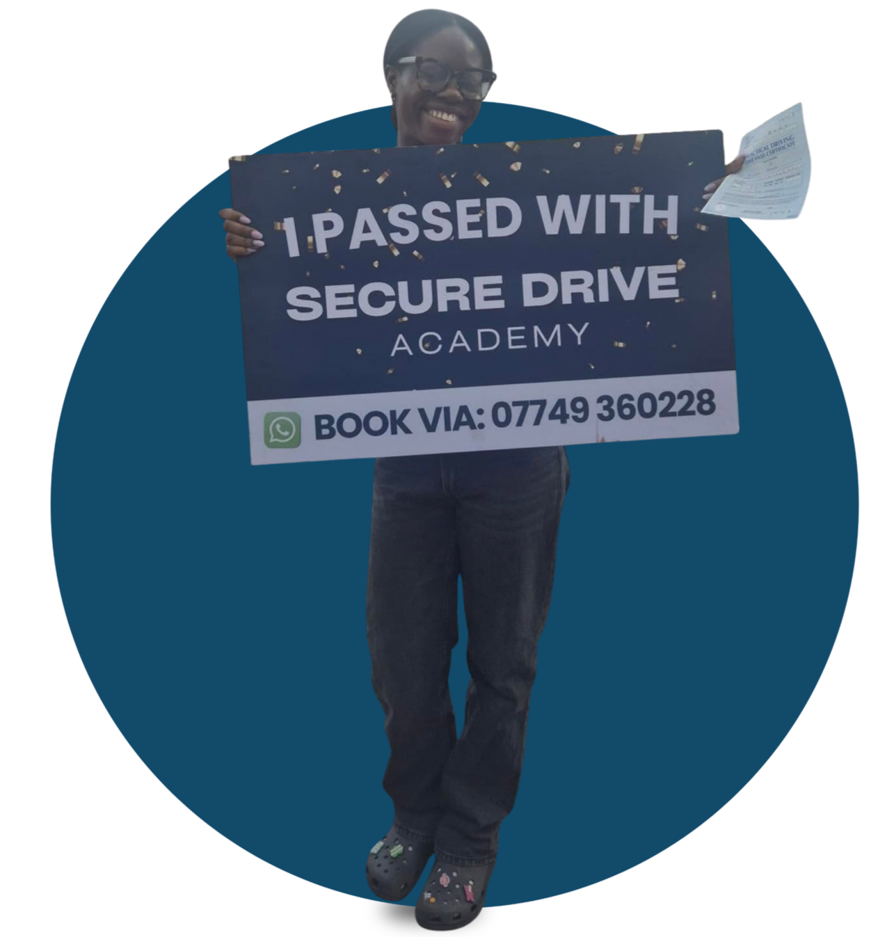 Person holding a sign that reads "Passed with Secure Drive Academy" and a WhatsApp contact number, smiling and wearing glasses, dark jeans, and Crocs with colorful designs.