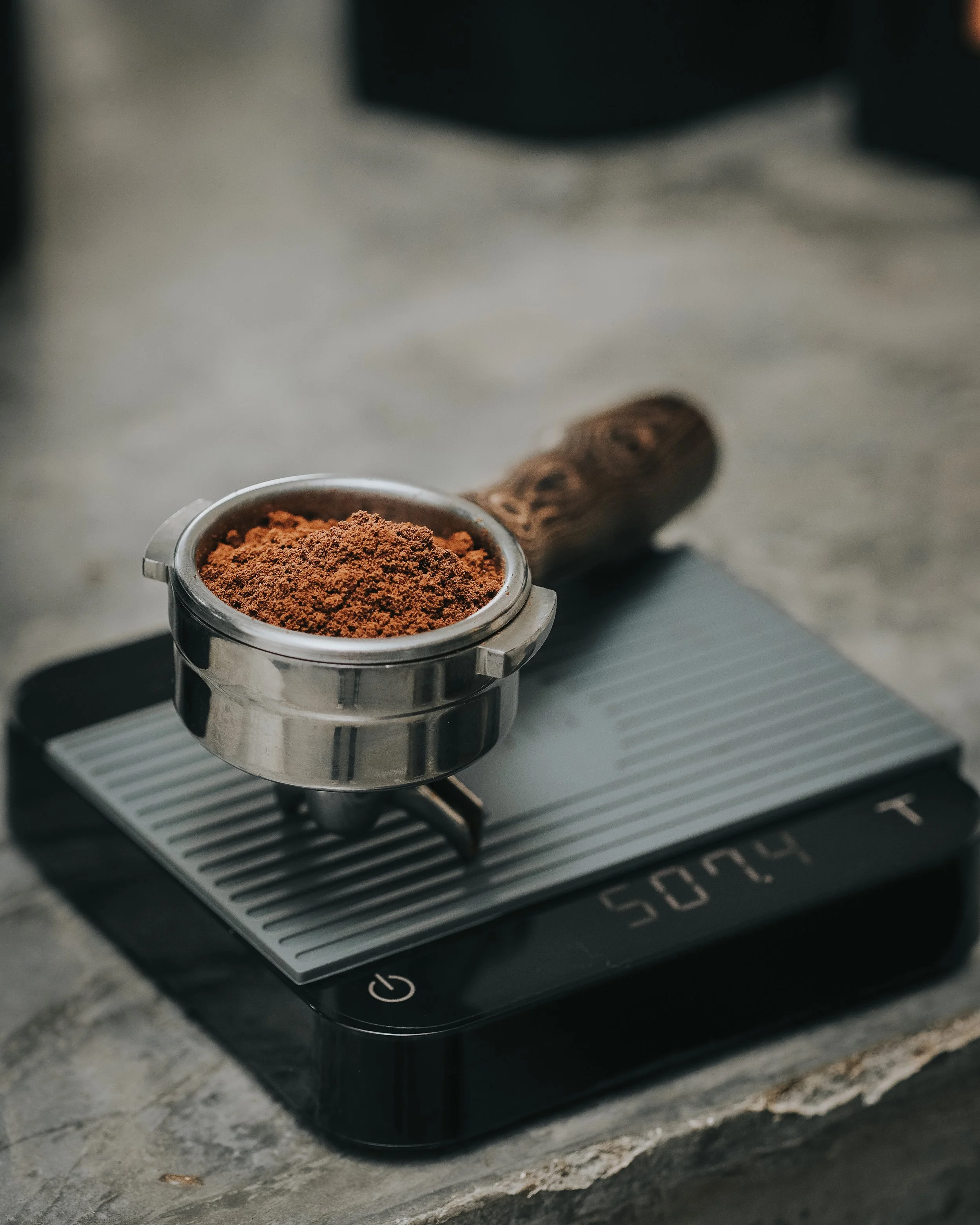 A portafilter filled with ground coffee resting on a digital coffee scale on a kitchen countertop.