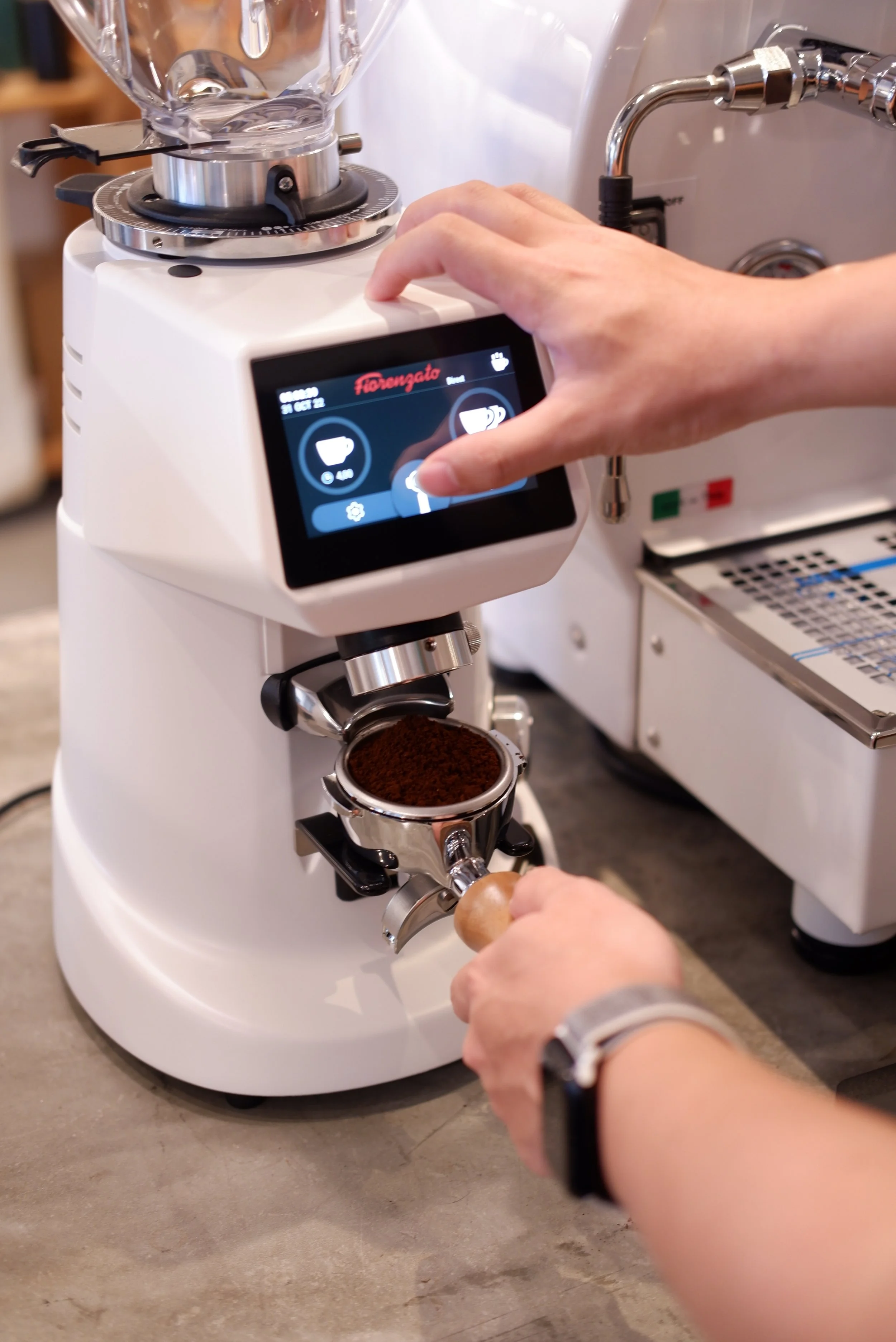 A person operating a coffee roasting machine, adjusting the controls with one hand and holding a wooden stick with the other, with ground coffee inside the machine.