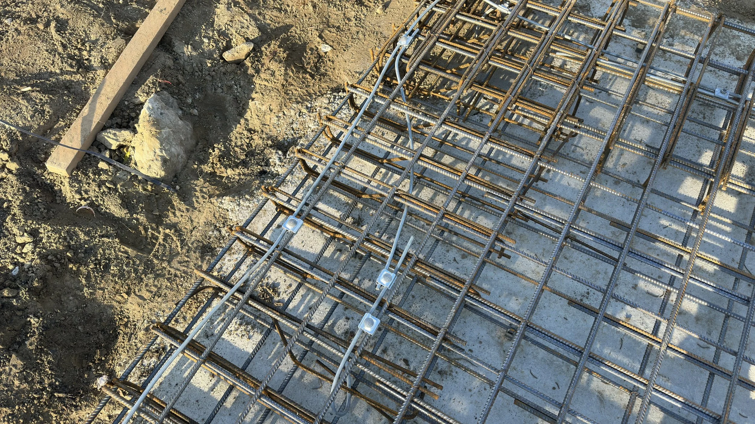 Építkezési terület alapozási fázisa, ahol a beton aljzatra helyezett acél vasrács háló látható, a háttérben földmunkával és doka zsaluzattal. Földelő szonda kiépítés látható. A földelés össze van kötve az alapvasalattal.
