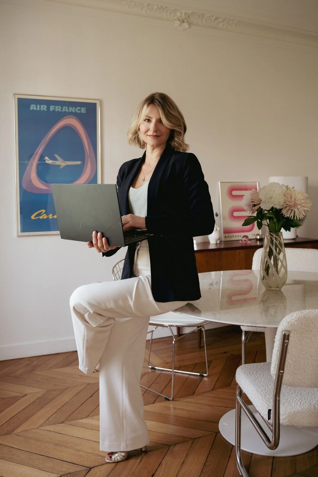 A woman standing in a room holding a laptop, with a framed poster of an airplane and 'Air France' on the wall behind her, and a vase of flowers on a table nearby.