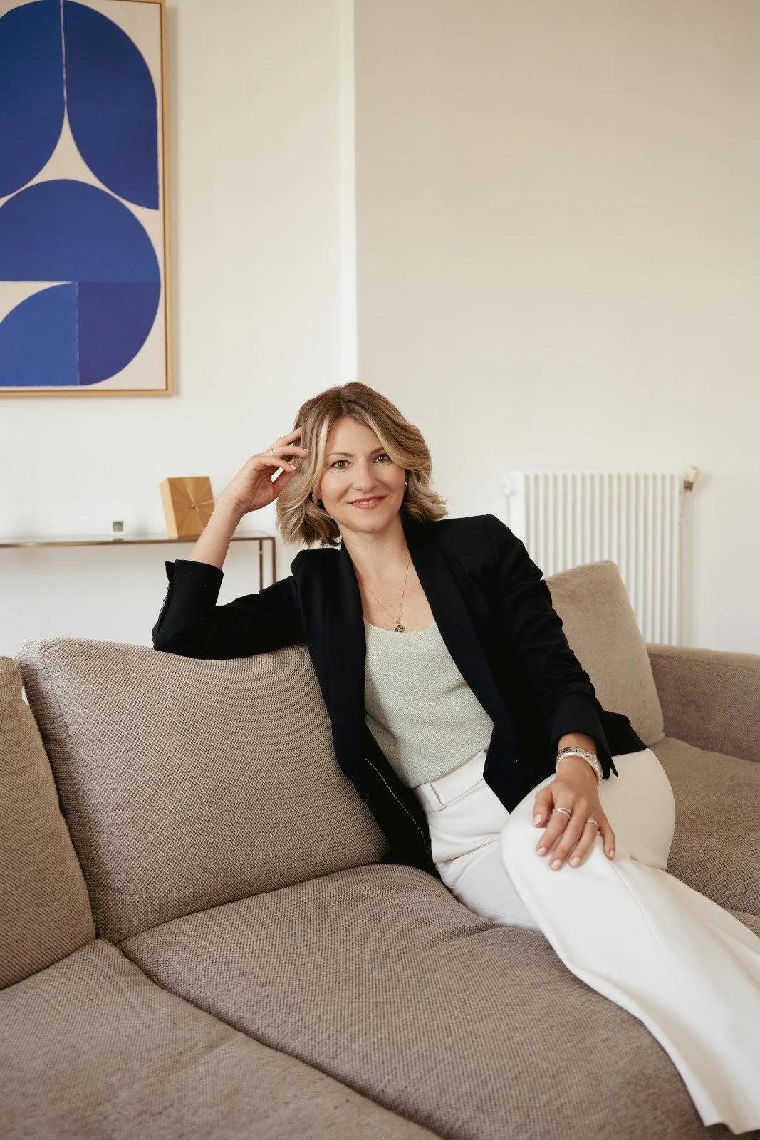 A woman with blonde hair sitting on a beige couch in a living room, smiling at the camera, wearing a black blazer and white pants.