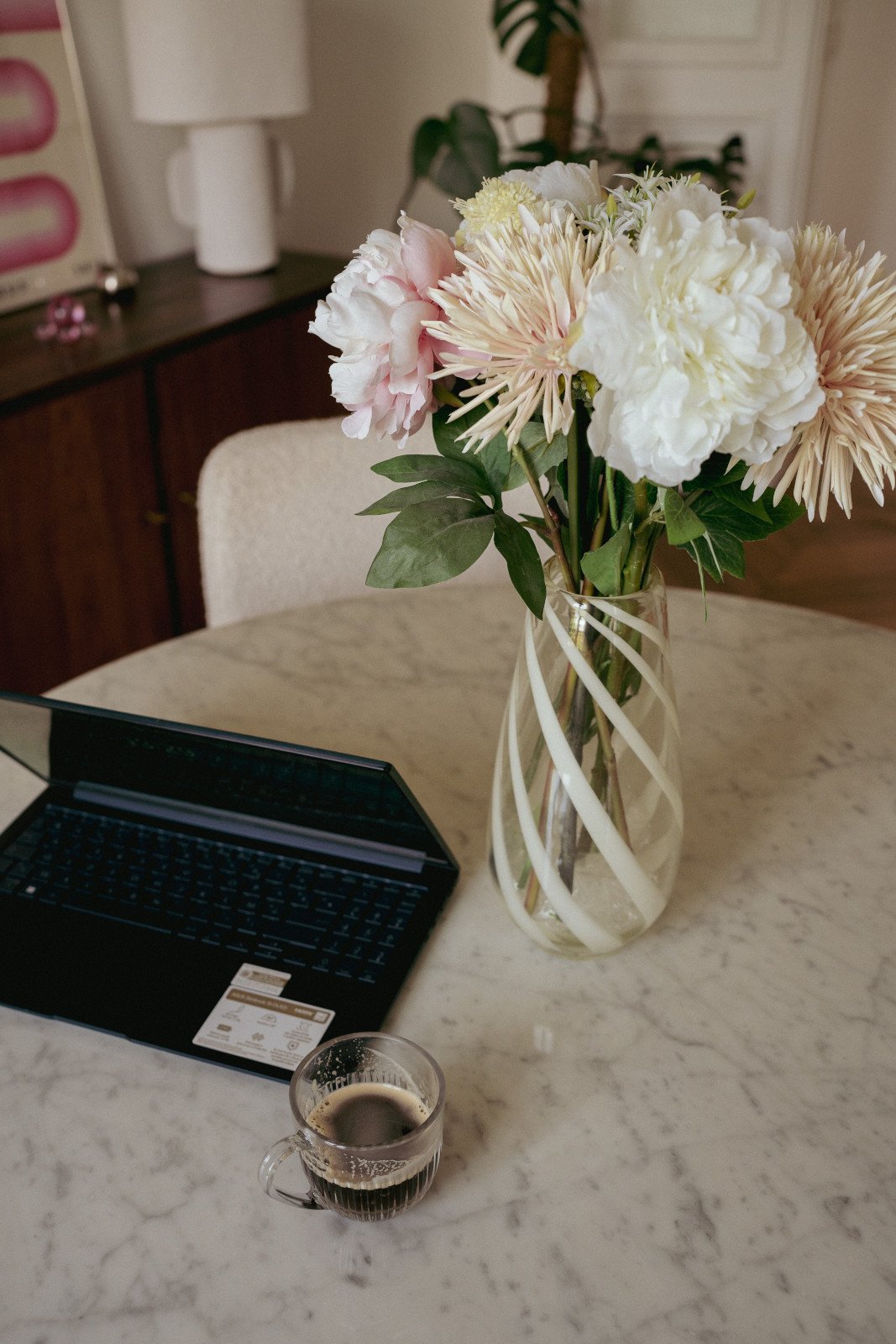 A vase with white and pink flowers and green leaves on a marble table, with a small glass of coffee, a laptop, a chair, a lamp, and a wooden cabinet in the background.