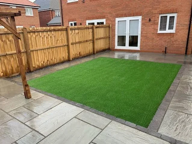 A backyard with a small patch of green artificial grass surrounded by gray stone pavers, enclosed by a wooden fence with a brick house in the background.